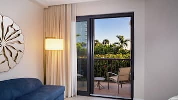 Habitación de hotel con sofá azul, lámpara, puerta de vidrio que da a un balcón con una silla y una mesa, y un adorno en la pared.