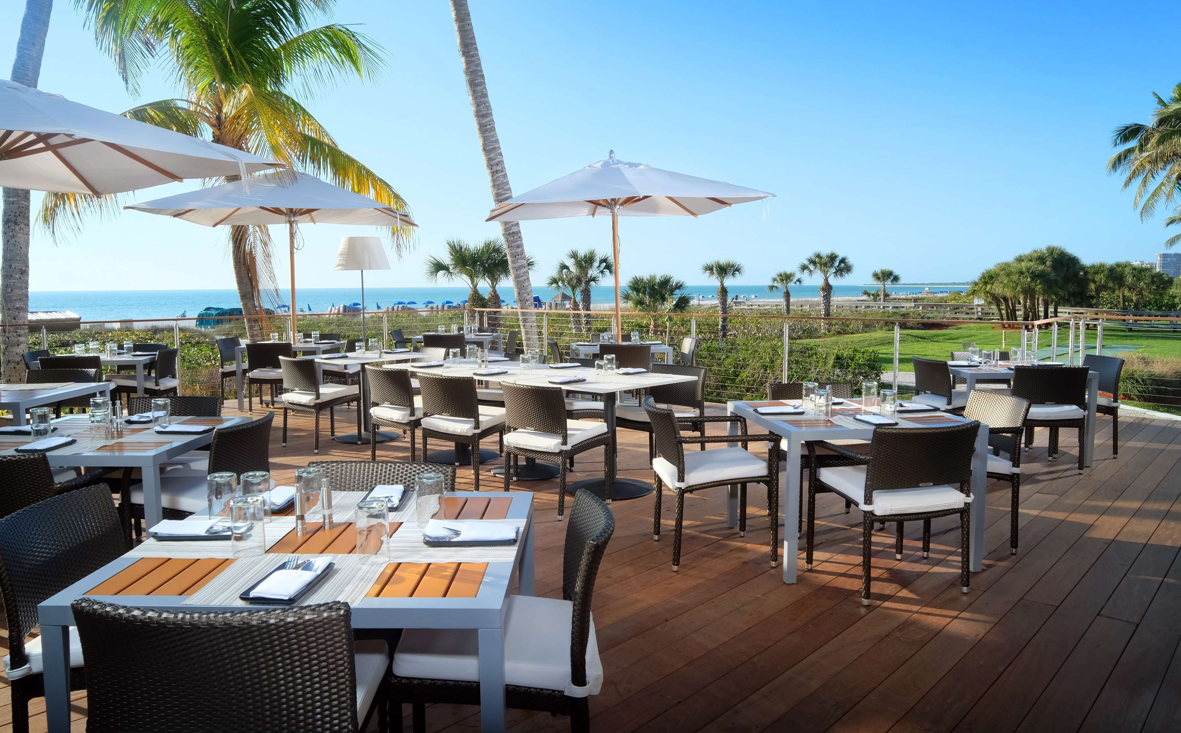 An outdoor dining area with tables and chairs, umbrellas, and a view of the ocean.