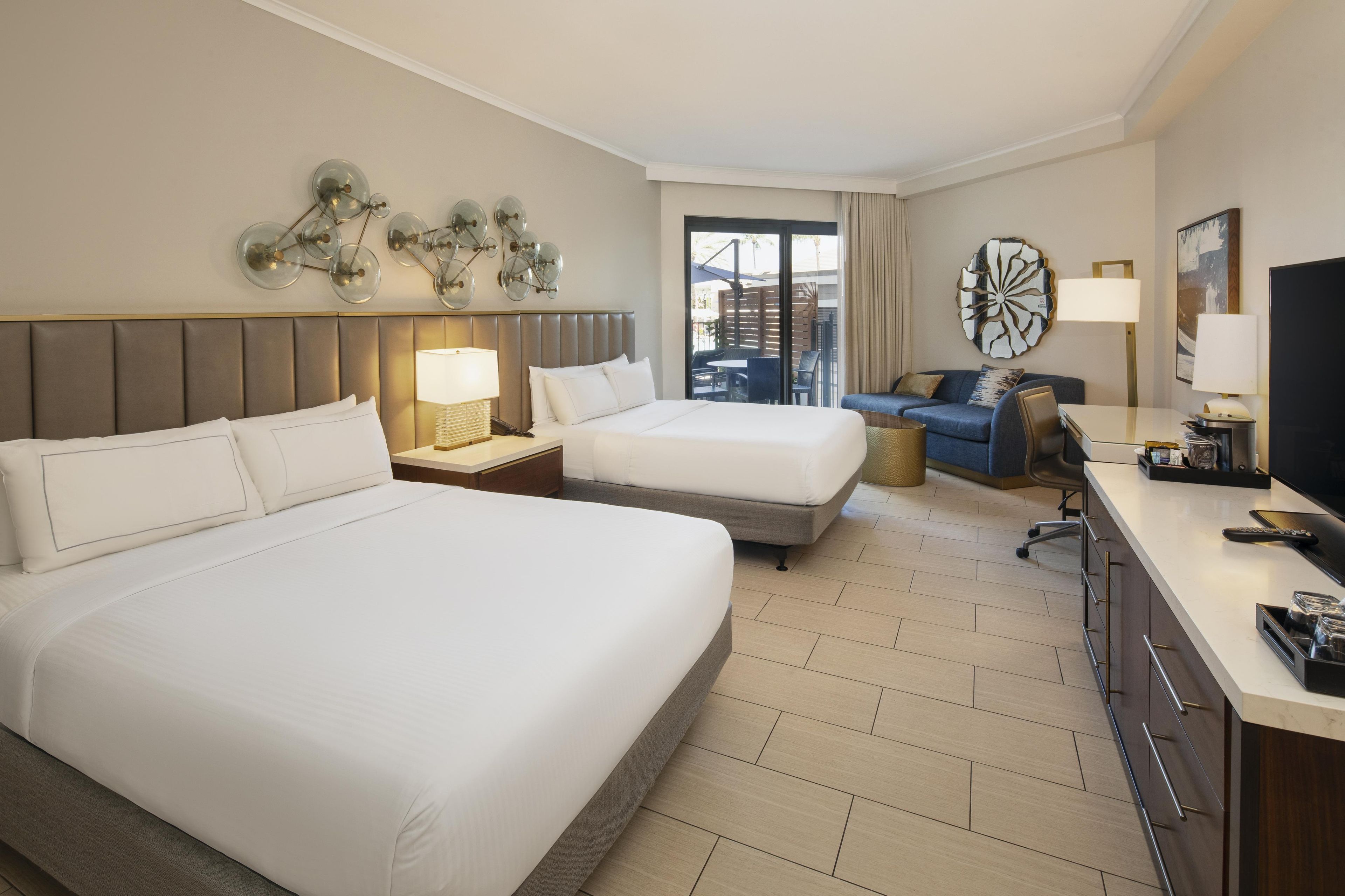 A hotel room with two beds, a desk, a television, and a view of the outdoors.