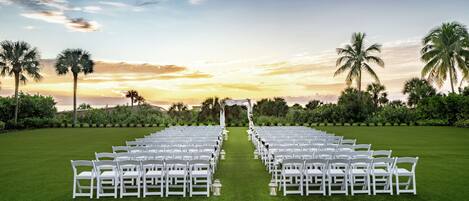 Une cérémonie de mariage est mise en place : des chaises blanches sont disposées en rangées sur une pelouse verdoyante, une arche blanche se dresse et des palmiers se trouvent en arrière-plan.