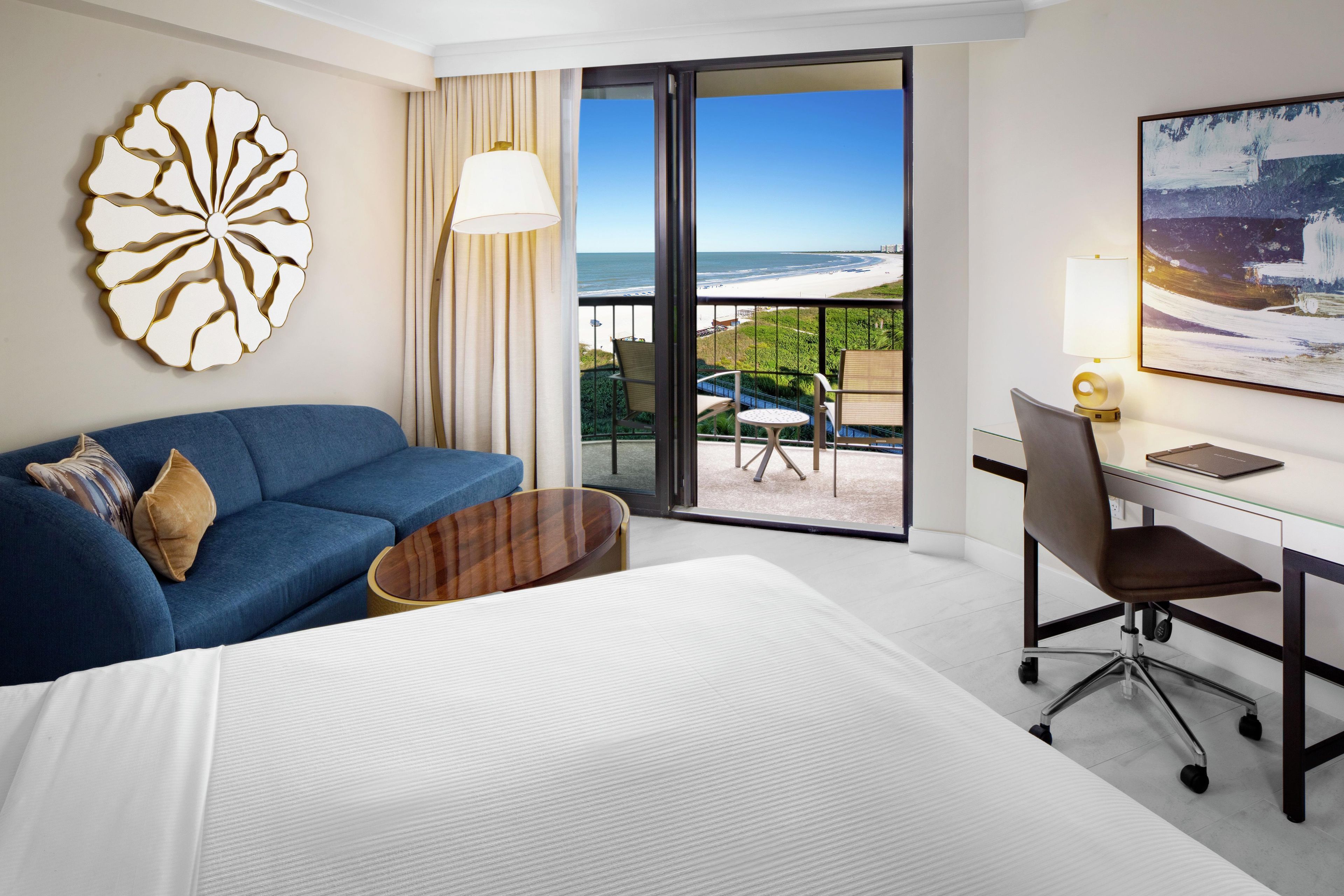 A hotel room with a bed, a blue sofa, a desk with a chair, a large window with a view of the beach, and a decorative wall piece.