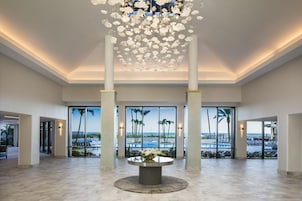A spacious lobby with a central round table, large windows with a view of palm trees and the sea, and a decorative hanging light fixture.