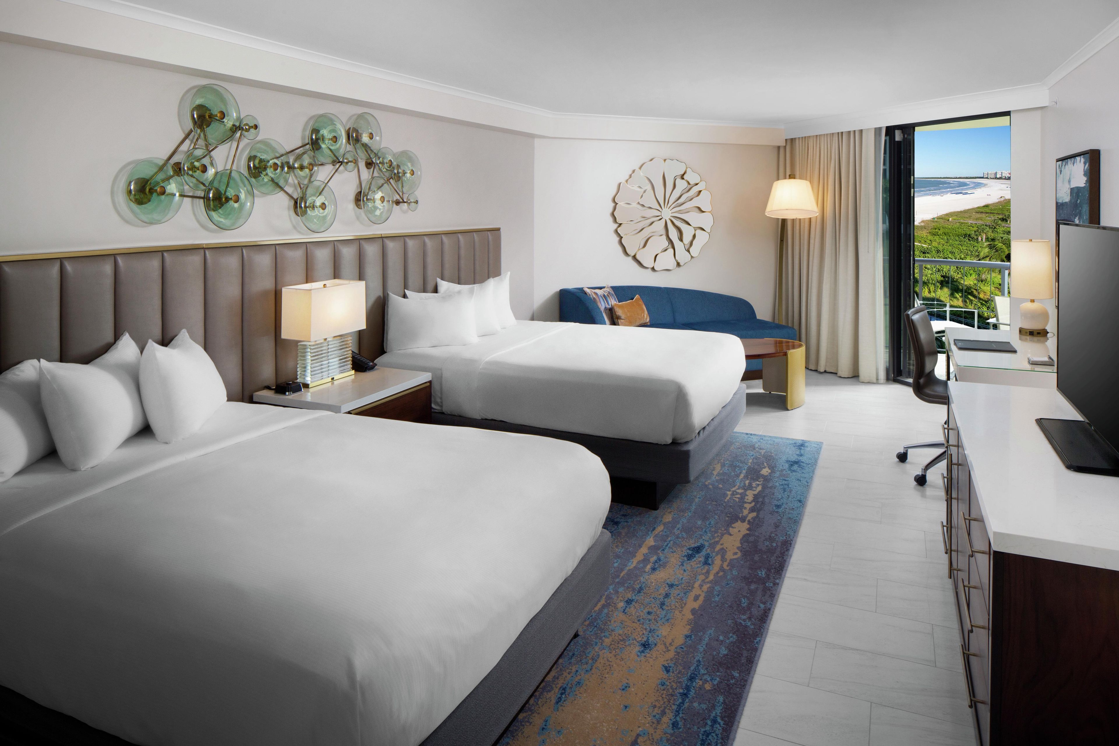 A hotel room with two beds, a desk, a chair, a TV, and a large window with a view of the beach.