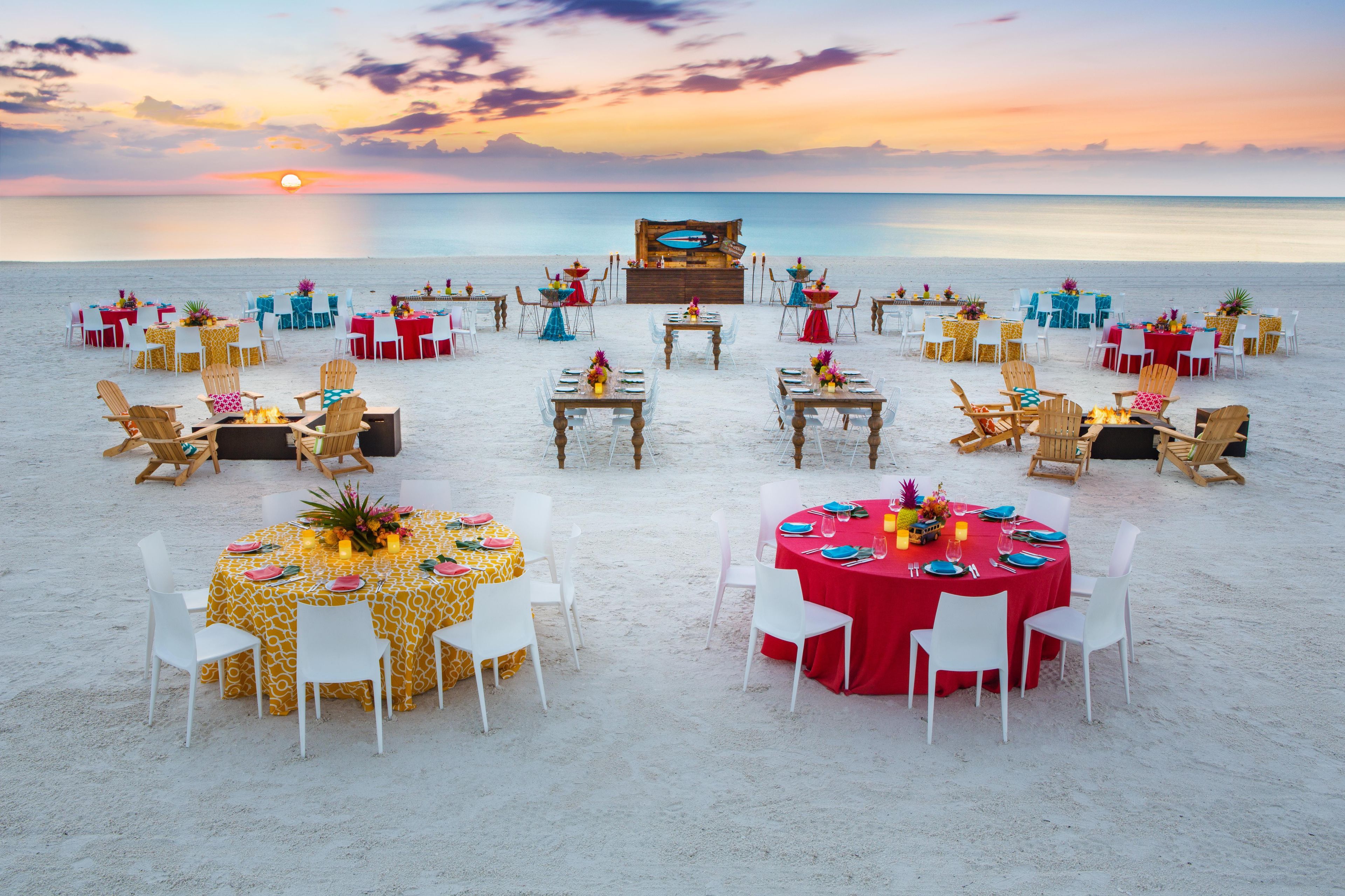 A beachside dining setup with multiple tables and chairs, arranged in a semi-circle facing the ocean.