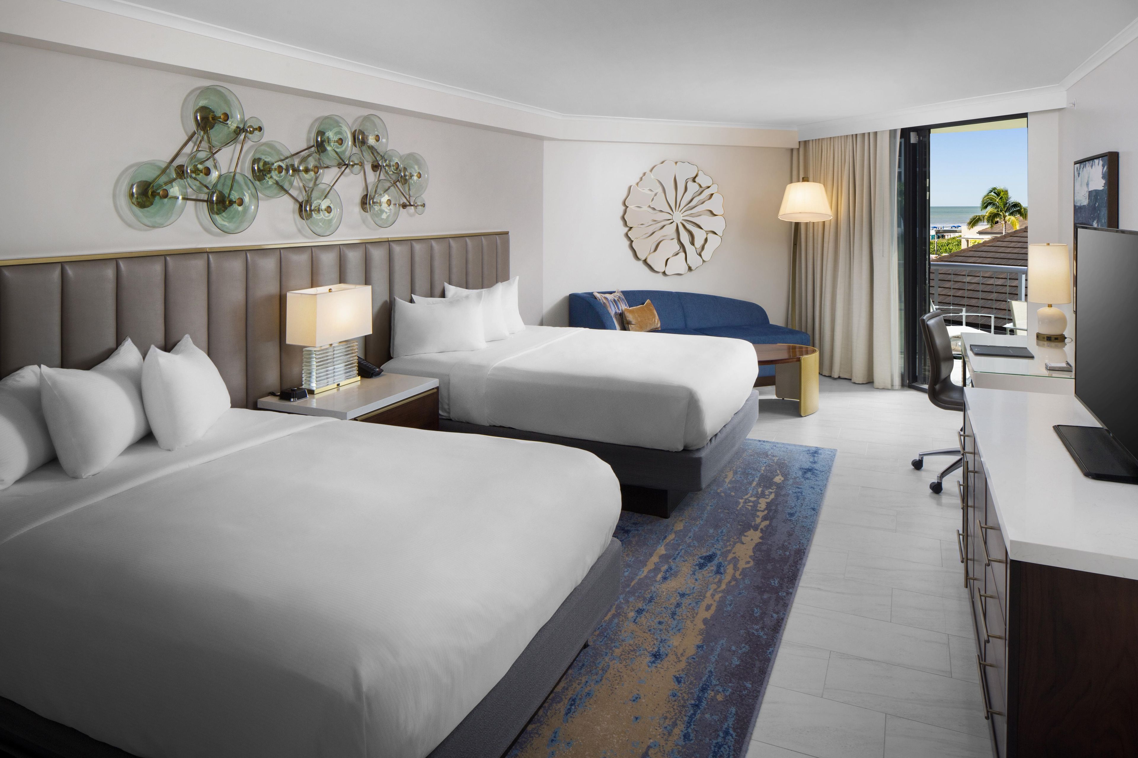 A hotel room with two beds, a desk, a chair, a television, and a view of the ocean.