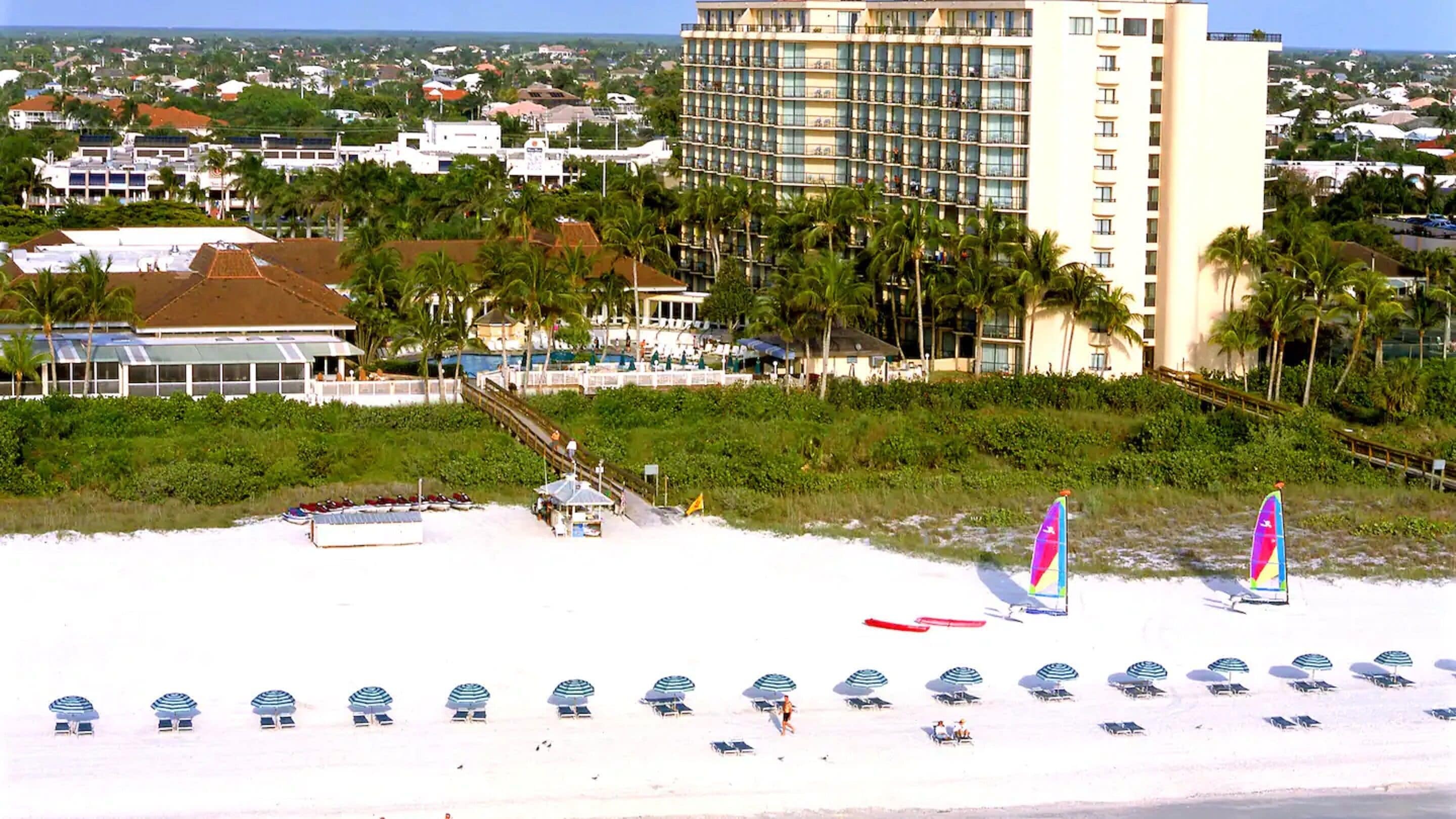 A beachfront hotel with multiple floors, a pool area, and numerous beach umbrellas.