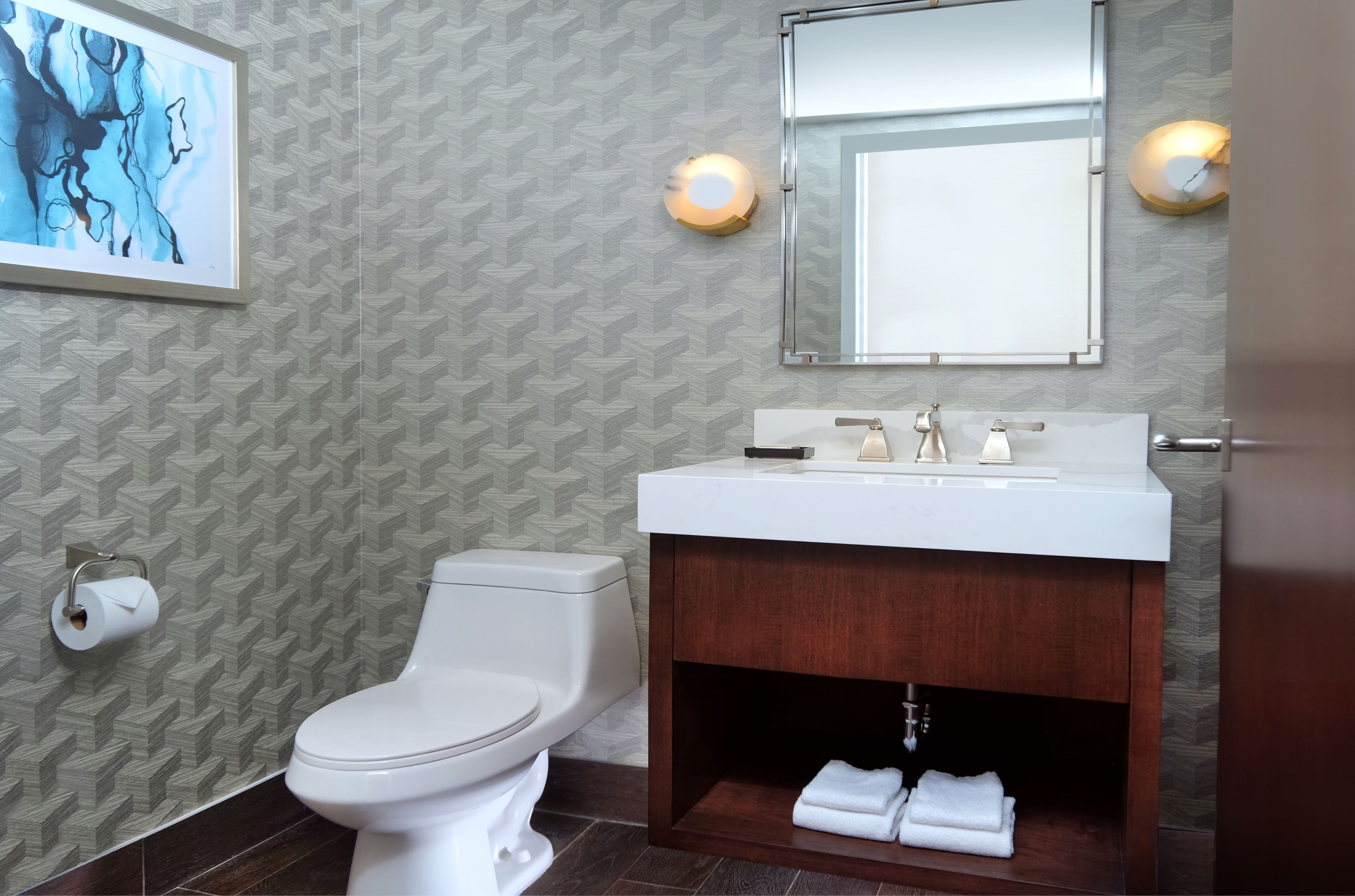 A modern bathroom with a toilet, sink, and mirror. There is a towel rack under the sink and a framed artwork on the wall.