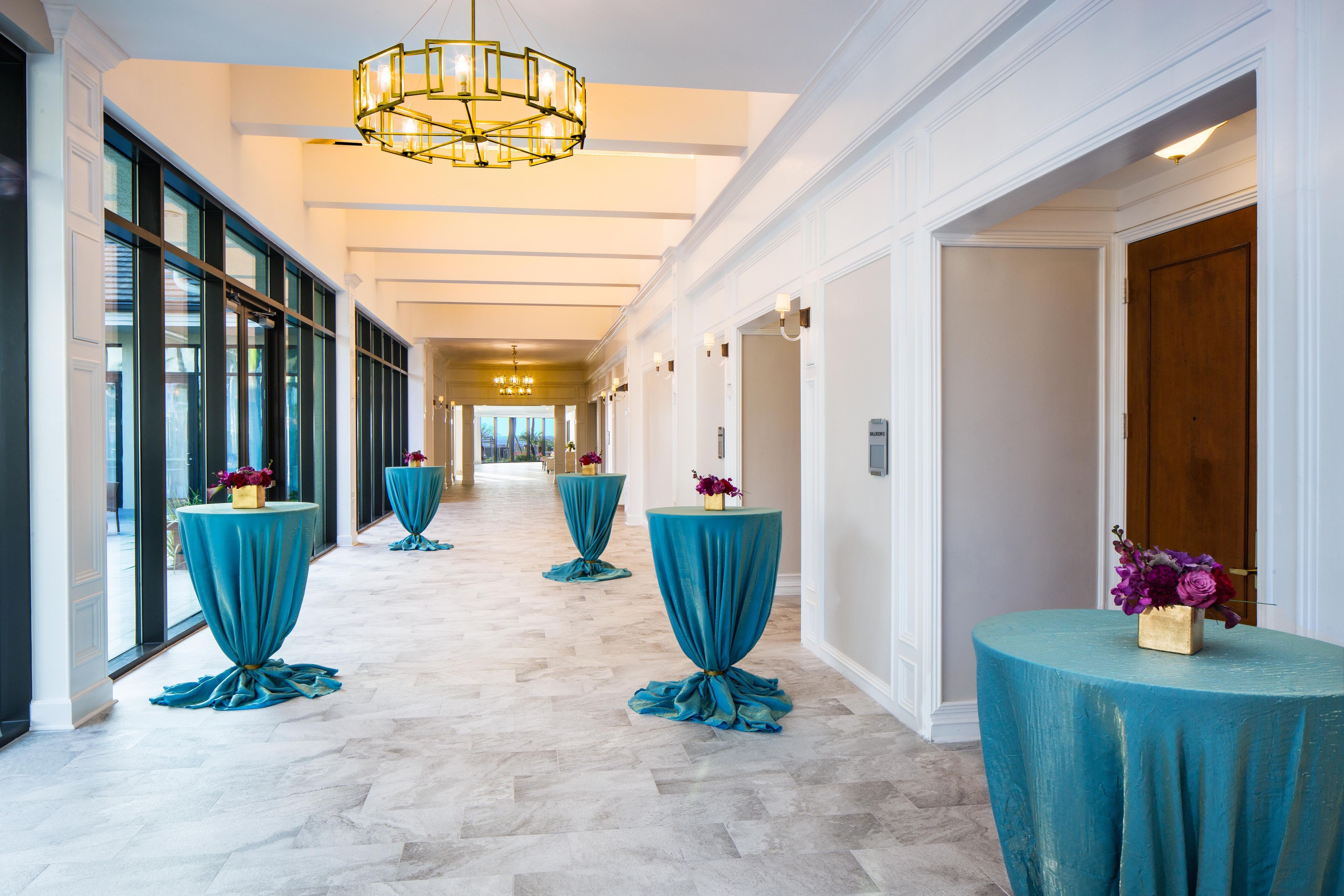 A hotel corridor with a marble floor, teal-covered tables with flowers, and a chandelier.