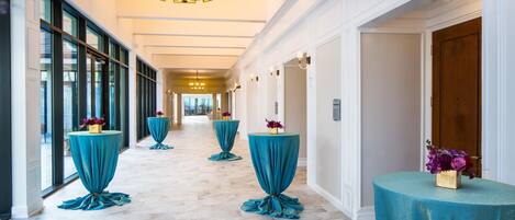 A hotel corridor with a marble floor, teal-covered tables with flowers, and a chandelier.