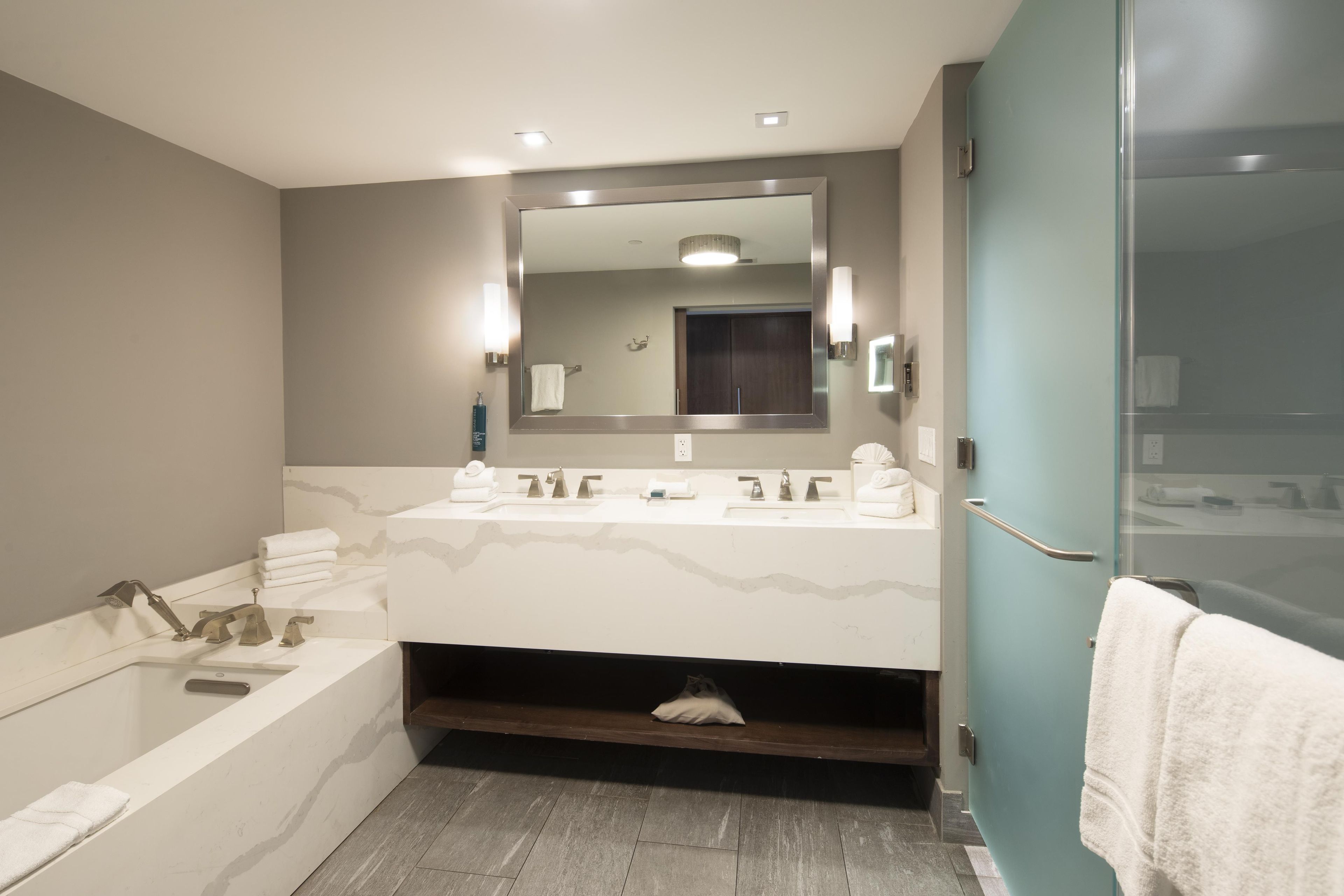 A modern bathroom with a large mirror, two sinks, a bathtub, and a shower area.