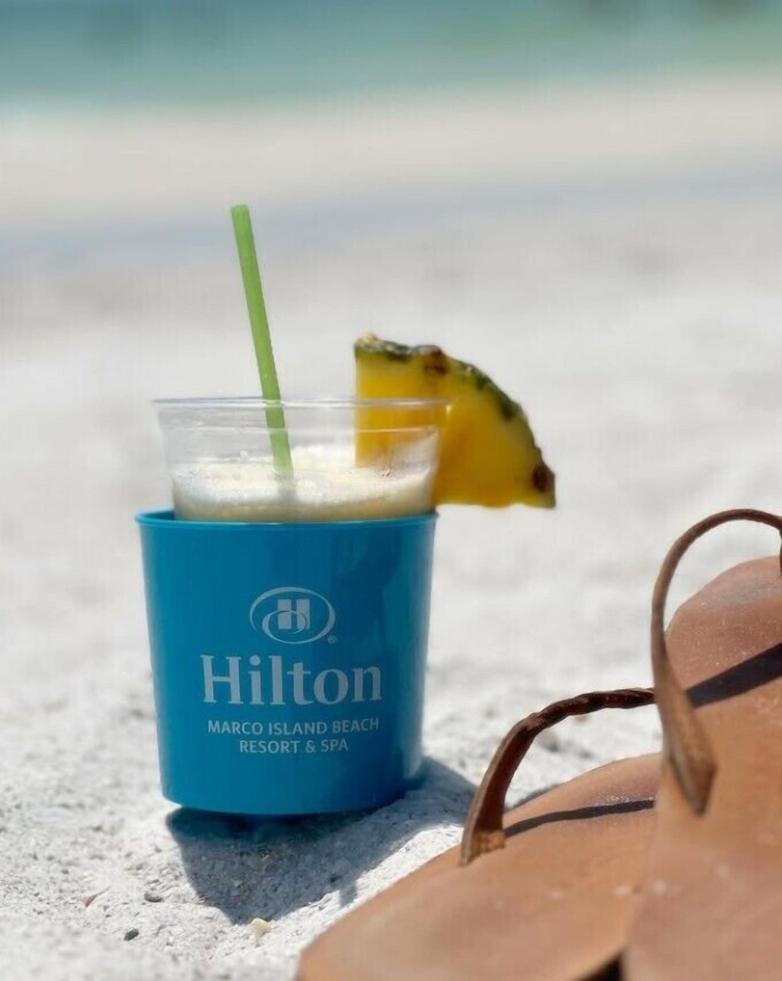 A blue Hilton branded drink cup with a straw and a slice of pineapple on a concrete surface.
