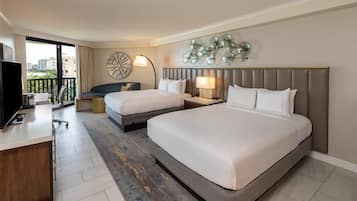 Habitación de hotel con dos camas, un escritorio, un televisor y un balcón con vista a la ciudad.