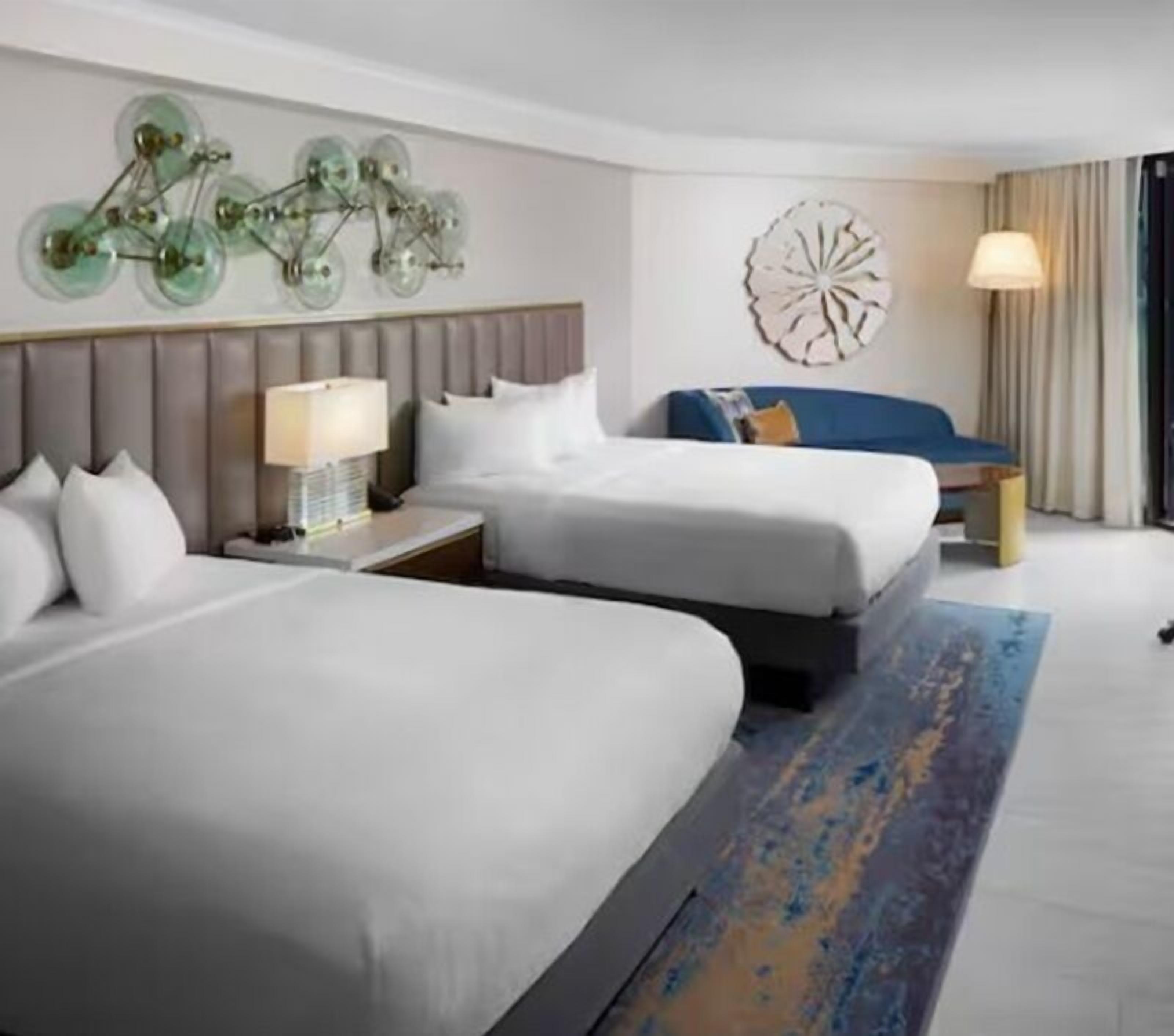 A hotel room with two beds, a nightstand with a lamp, a decorative wall piece, and a large window with curtains.