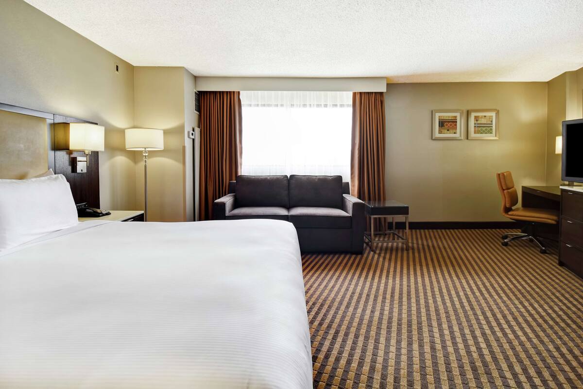 A hotel room with a large bed, a sofa, a desk, a chair, a television, and a window with curtains.