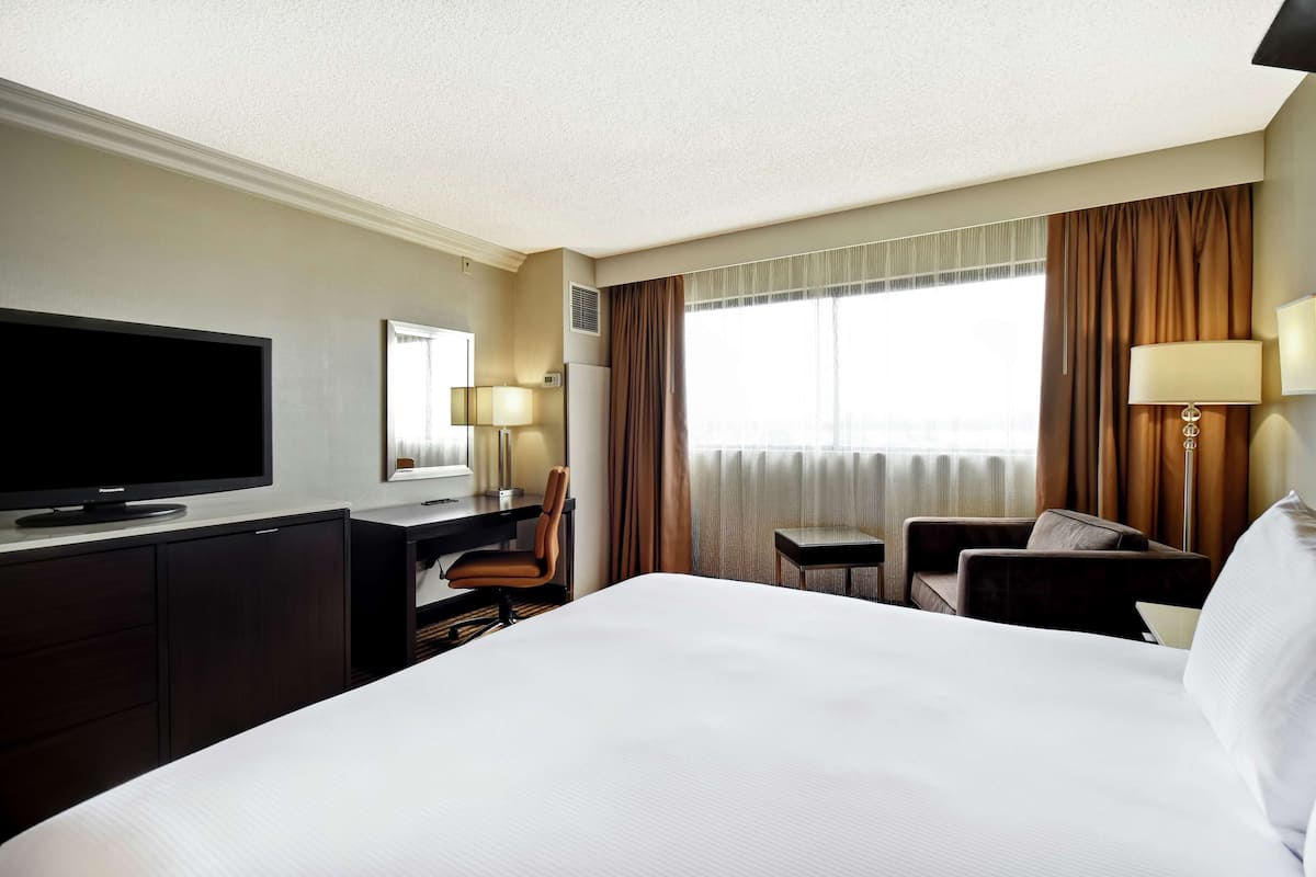 A hotel room with a large bed, a desk with a flat-screen TV, a chair, a small table, and a lamp.