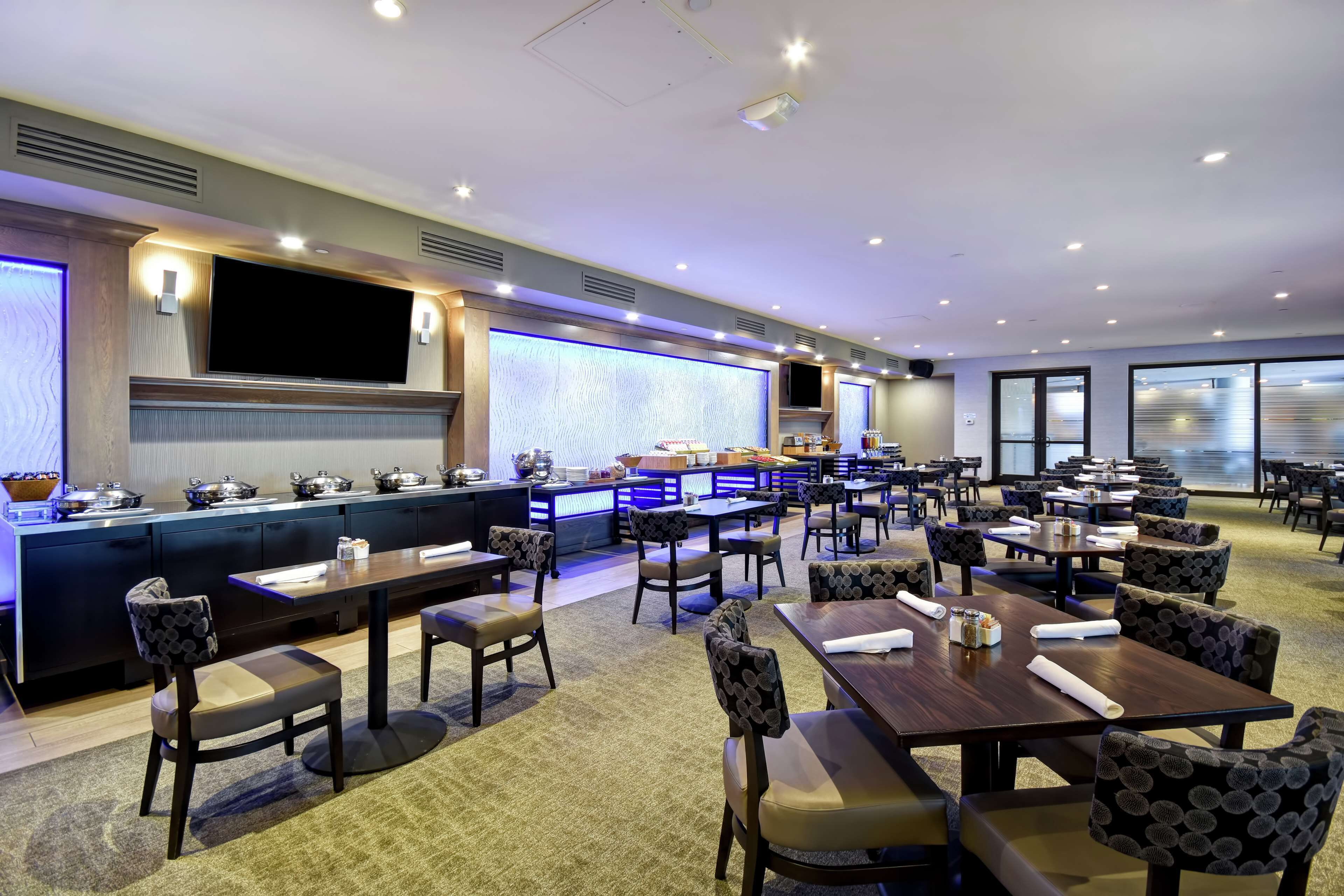 A hotel dining area with multiple tables and chairs, a kitchen counter, and a large flat-screen TV.