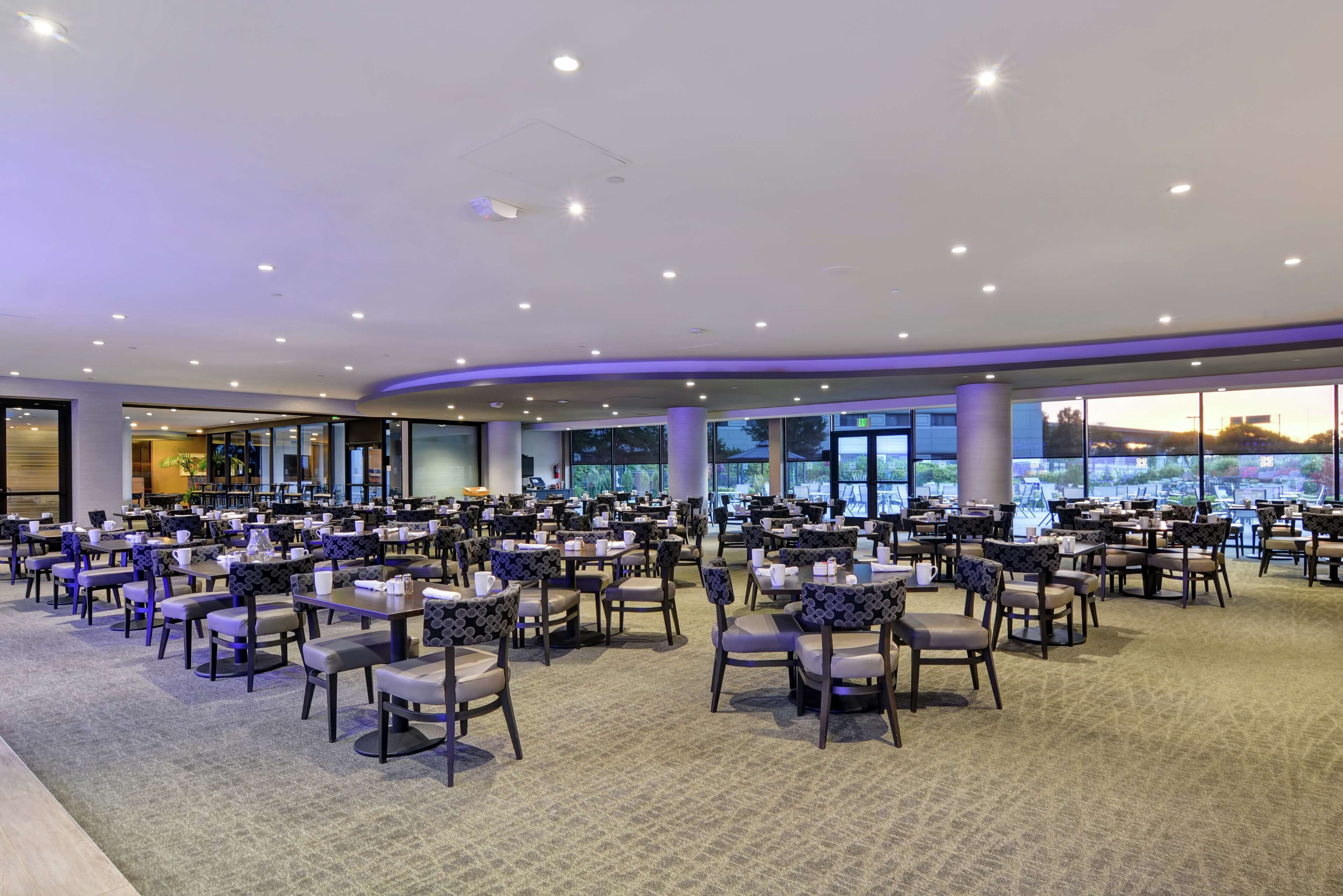 A spacious dining area with multiple tables and chairs, large windows offering a view, and a carpeted floor.
