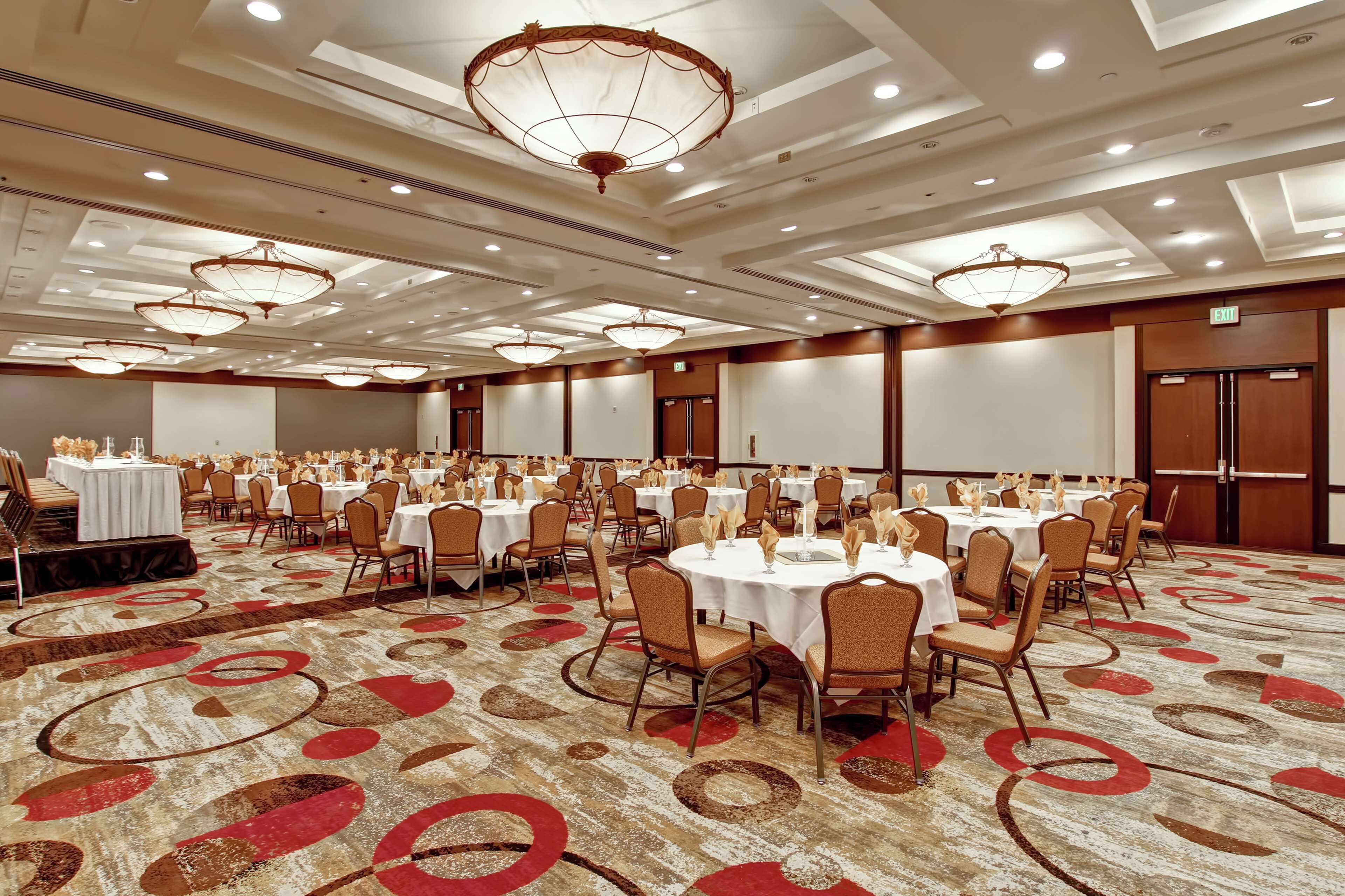 A large banquet hall with round tables and chairs, a stage, and a carpeted floor.