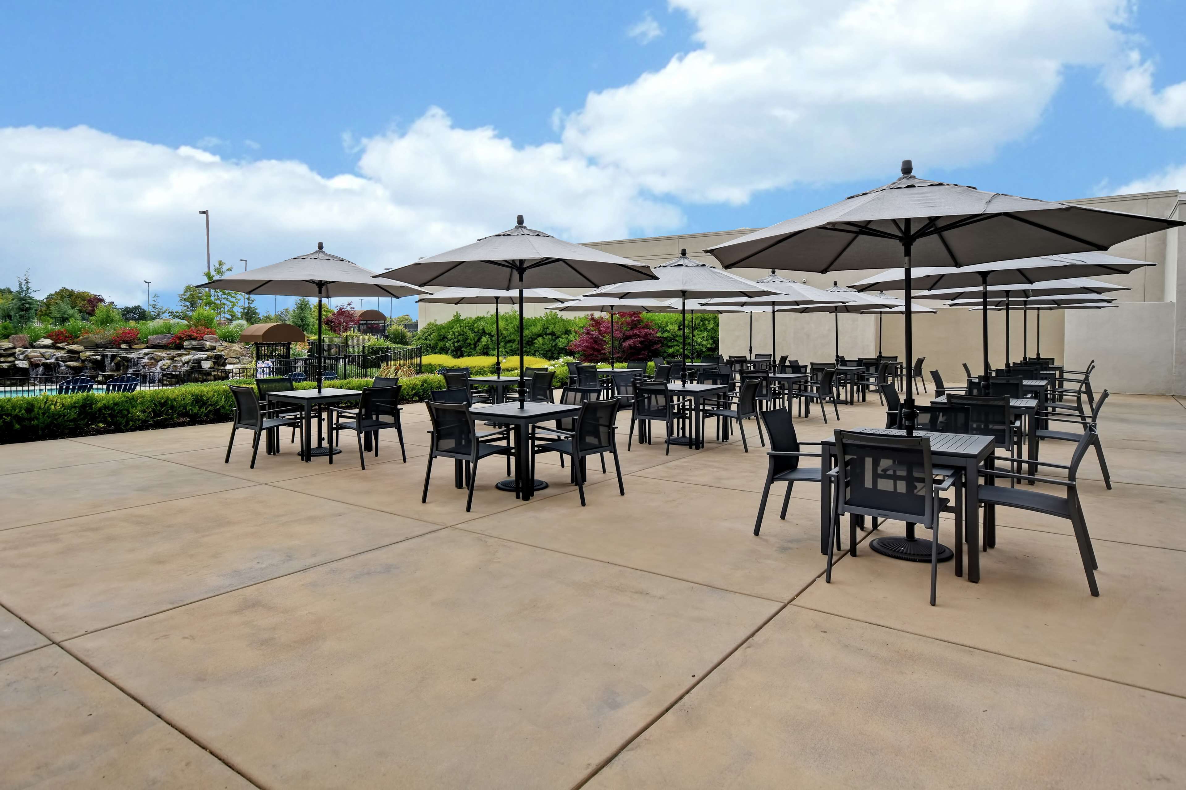 An outdoor patio with multiple tables and chairs, large umbrellas, and a pool in the background.