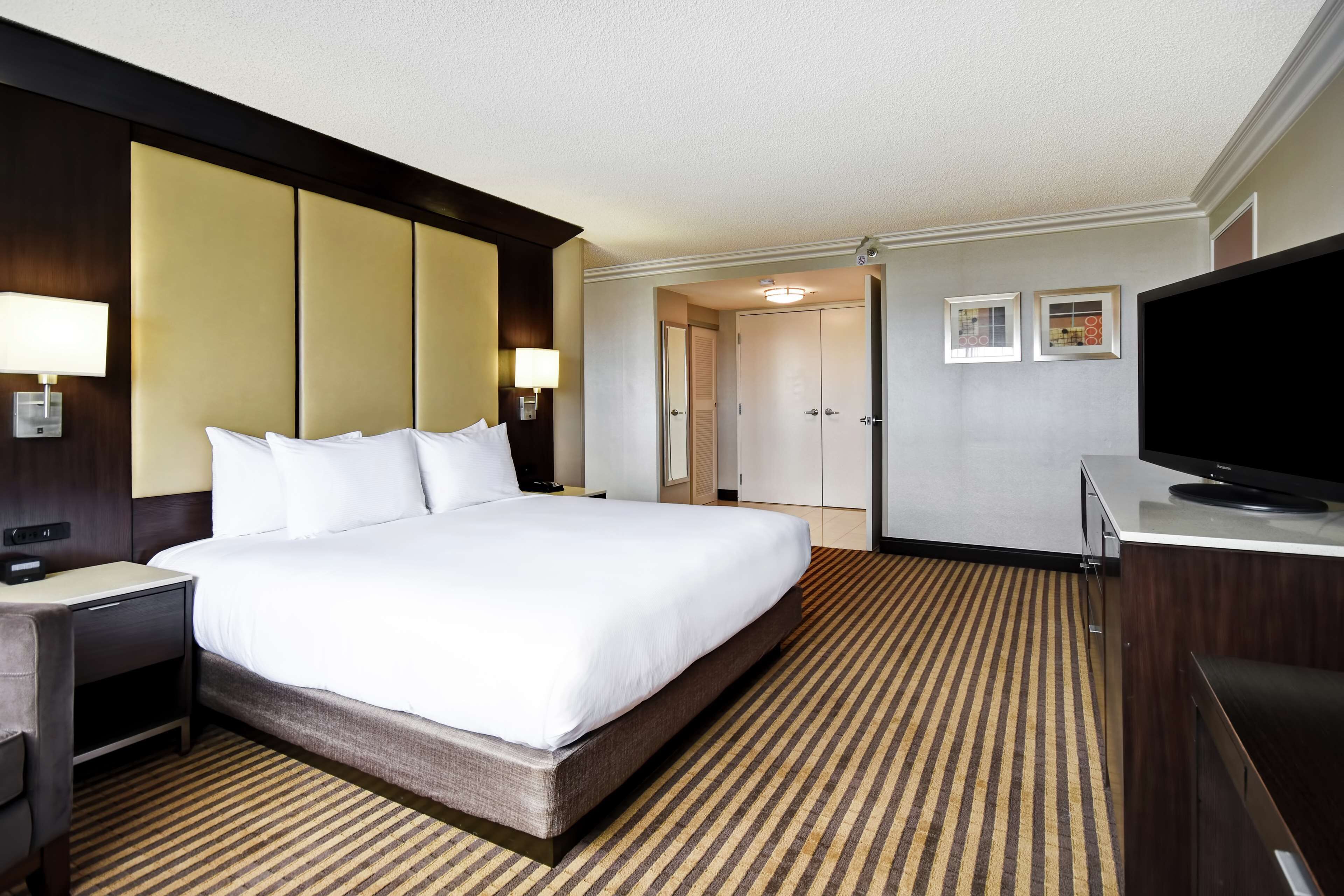 Quarto de hotel com uma cama grande, televisão, escrivaninha com luminária e armário.