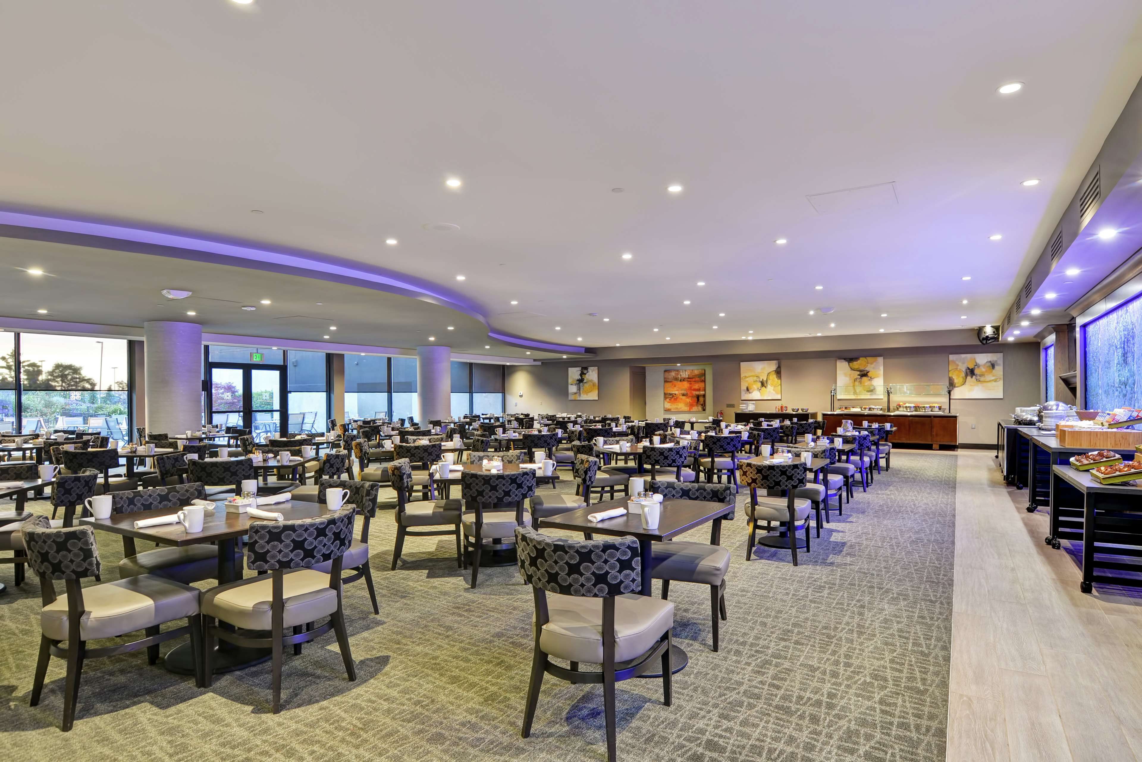 A spacious dining area with multiple tables and chairs, large windows, and a buffet setup.