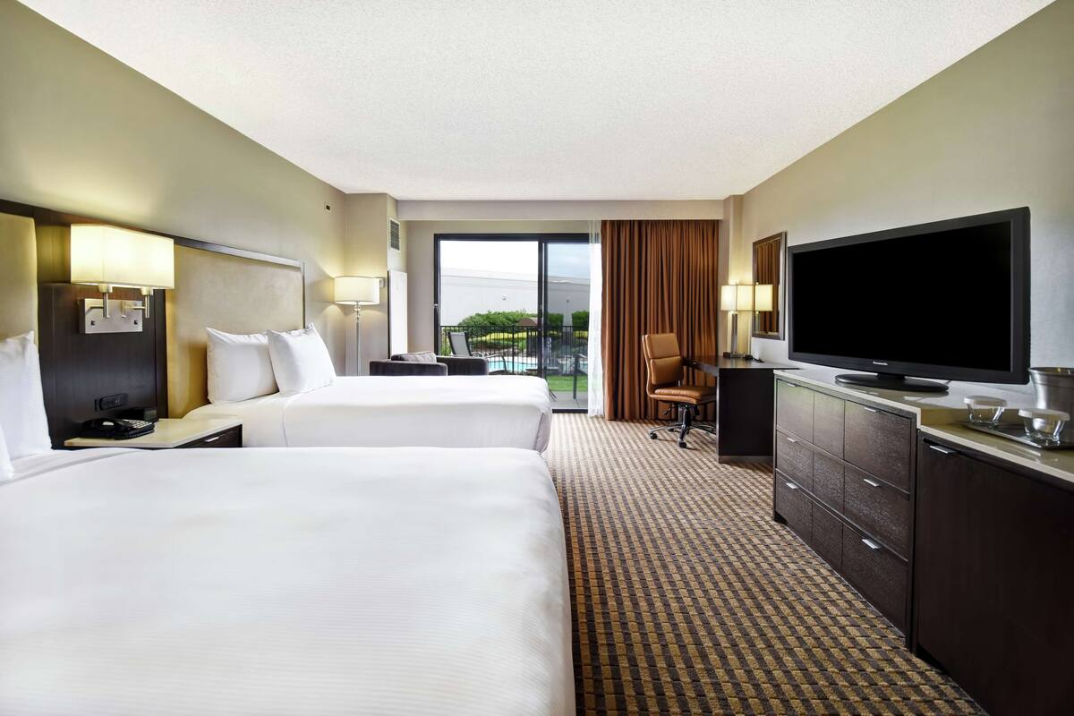 A hotel room with two beds, a flat-screen TV, a desk with a lamp, and a view of the outdoors.