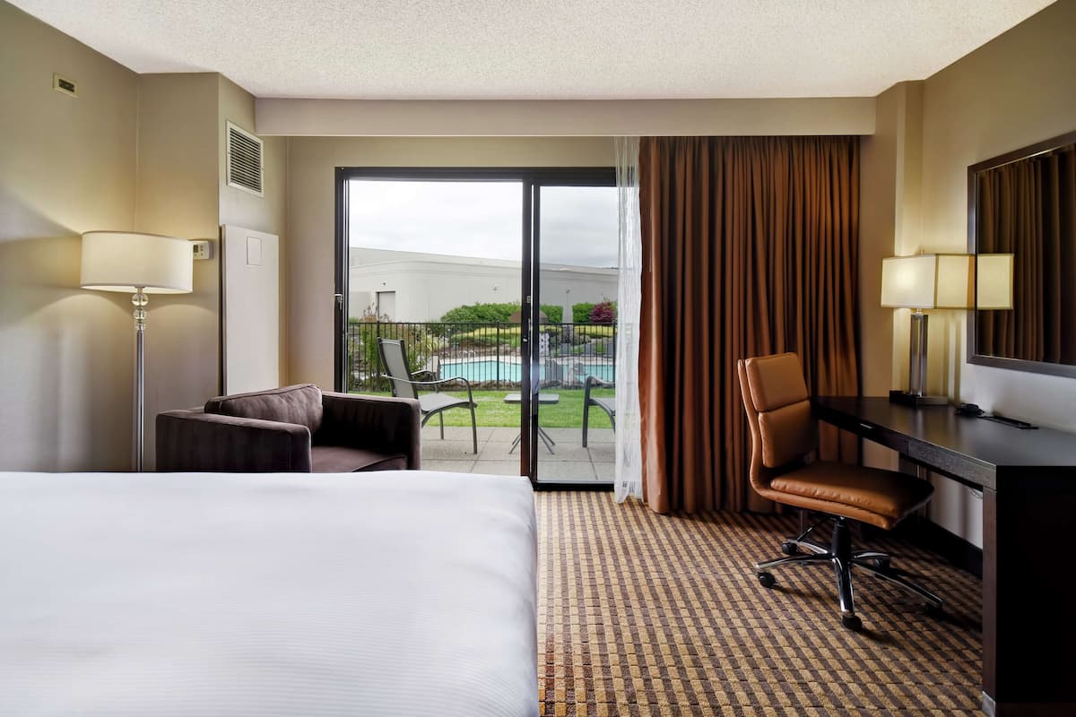 A hotel room with a large bed, a desk, a chair, and a view of a swimming pool through a sliding glass door.