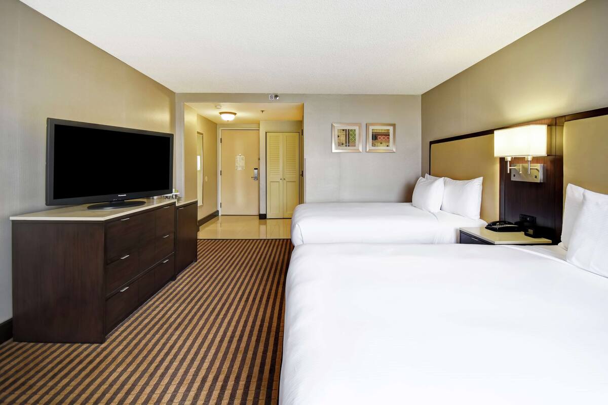 A hotel room with a large flat-screen TV, two beds, a dresser, and a carpeted floor.