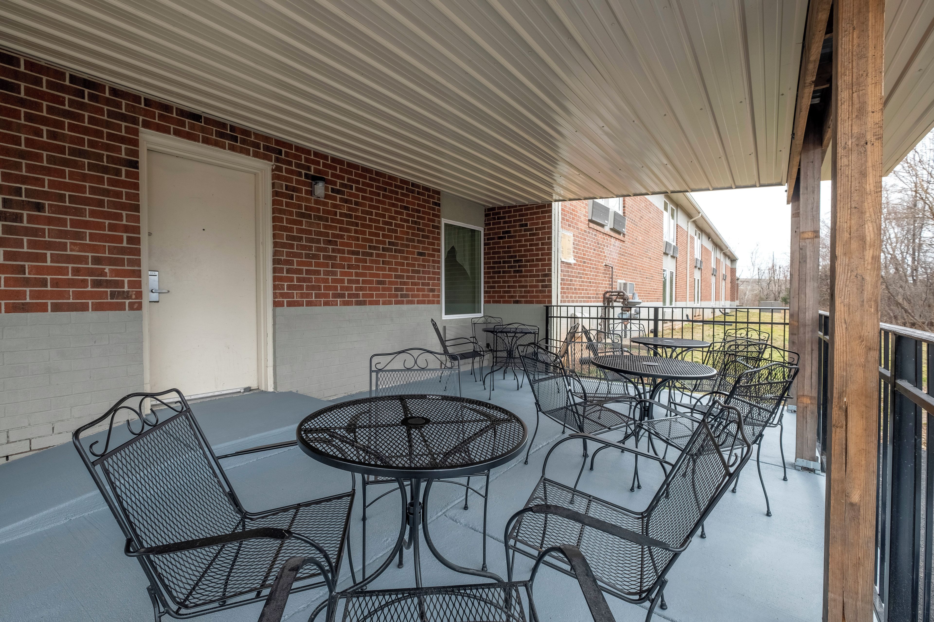 A covered outdoor patio with metal tables and chairs, a brick wall, and a door.