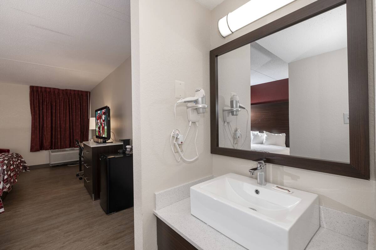 A hotel room with a large mirror, a white sink, a hairdryer, and a television on a dresser.
