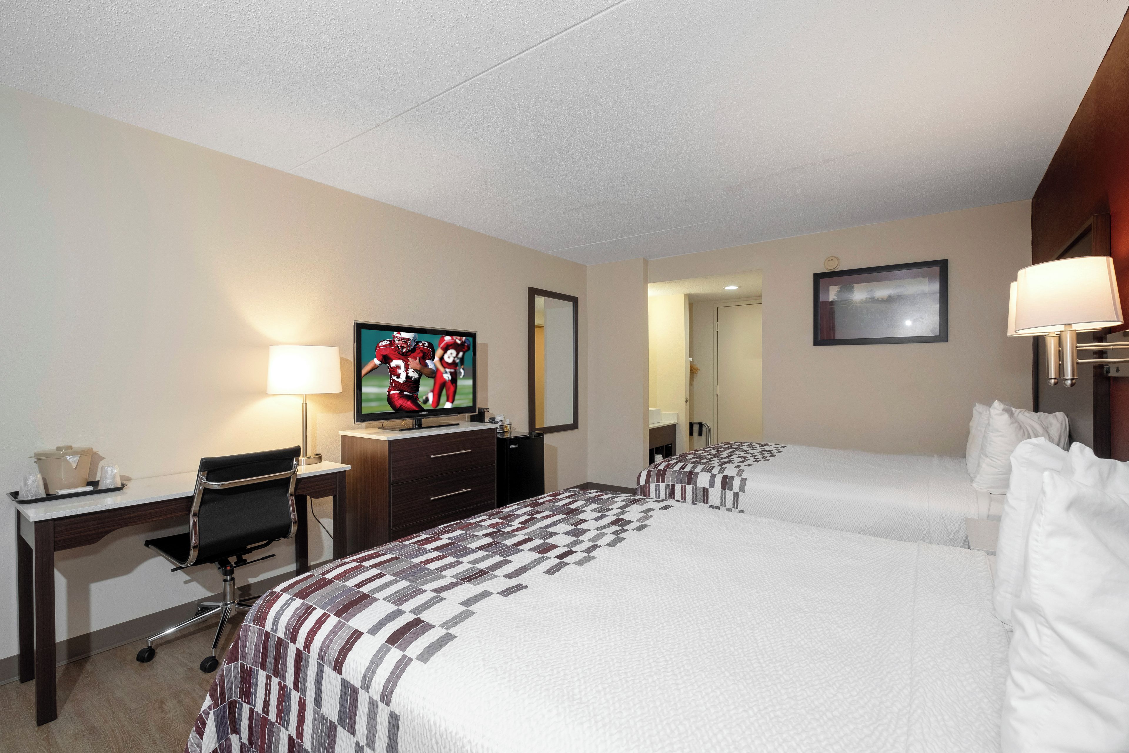 A hotel room with two beds, a desk with a chair, a television, a lamp, and a mirror.