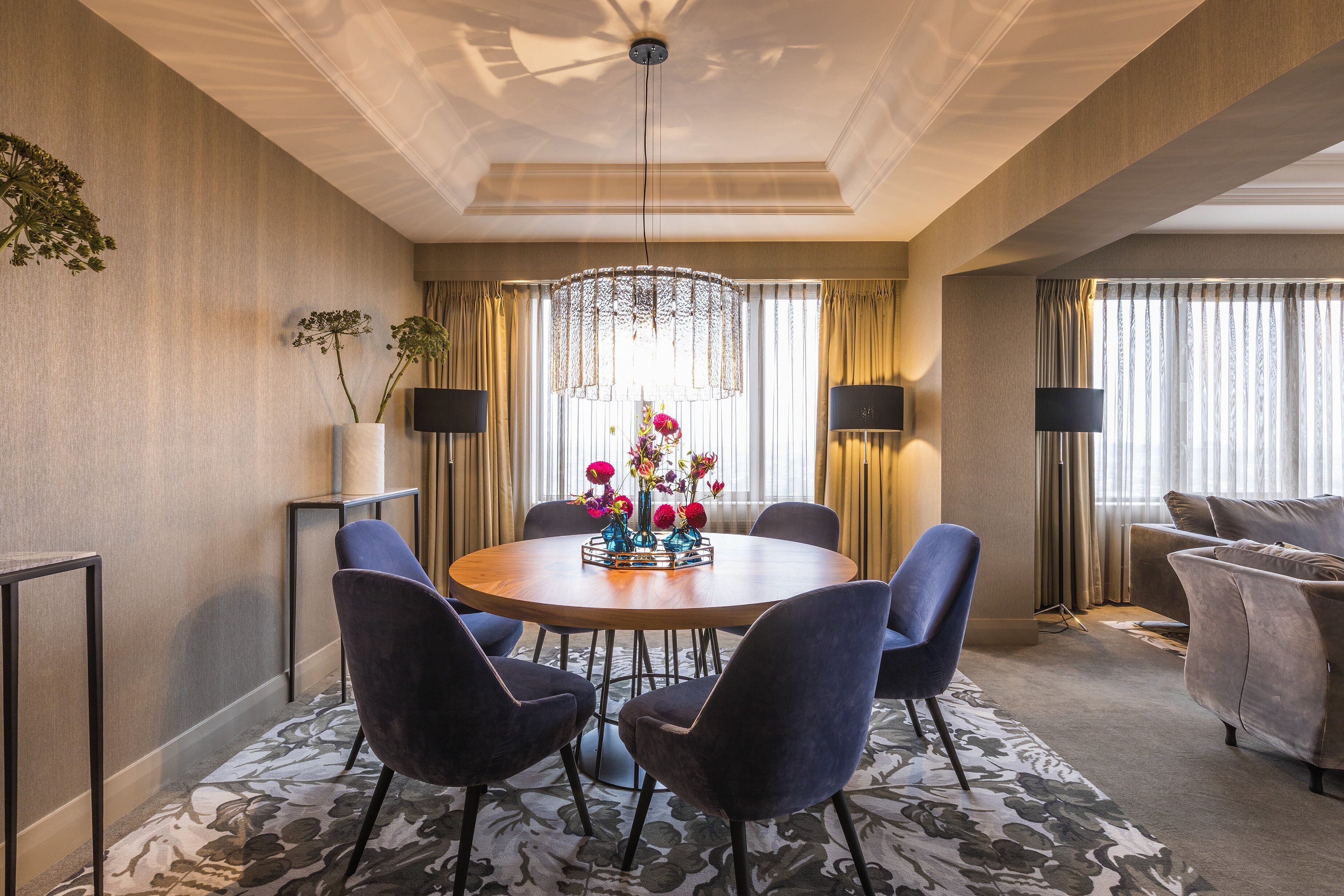 A modern dining area with a round table, six chairs, a chandelier, and a floral centerpiece.
