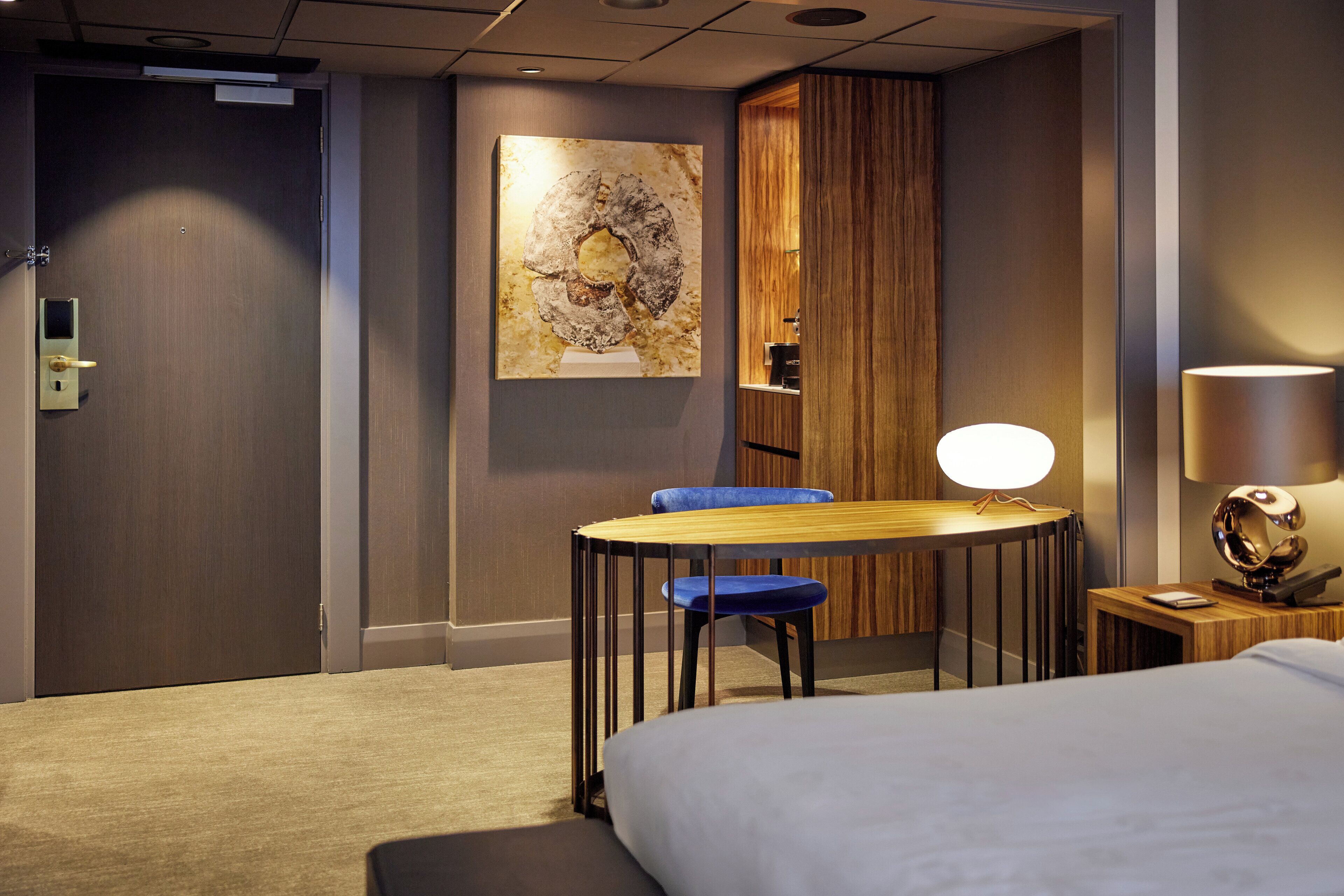 A hotel room with a bed, a desk with a chair, a lamp, and a painting on the wall.