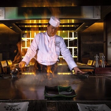 A chef cooking on a large, flat cooking surface in a modern kitchen with wooden paneling and stainless steel appliances.