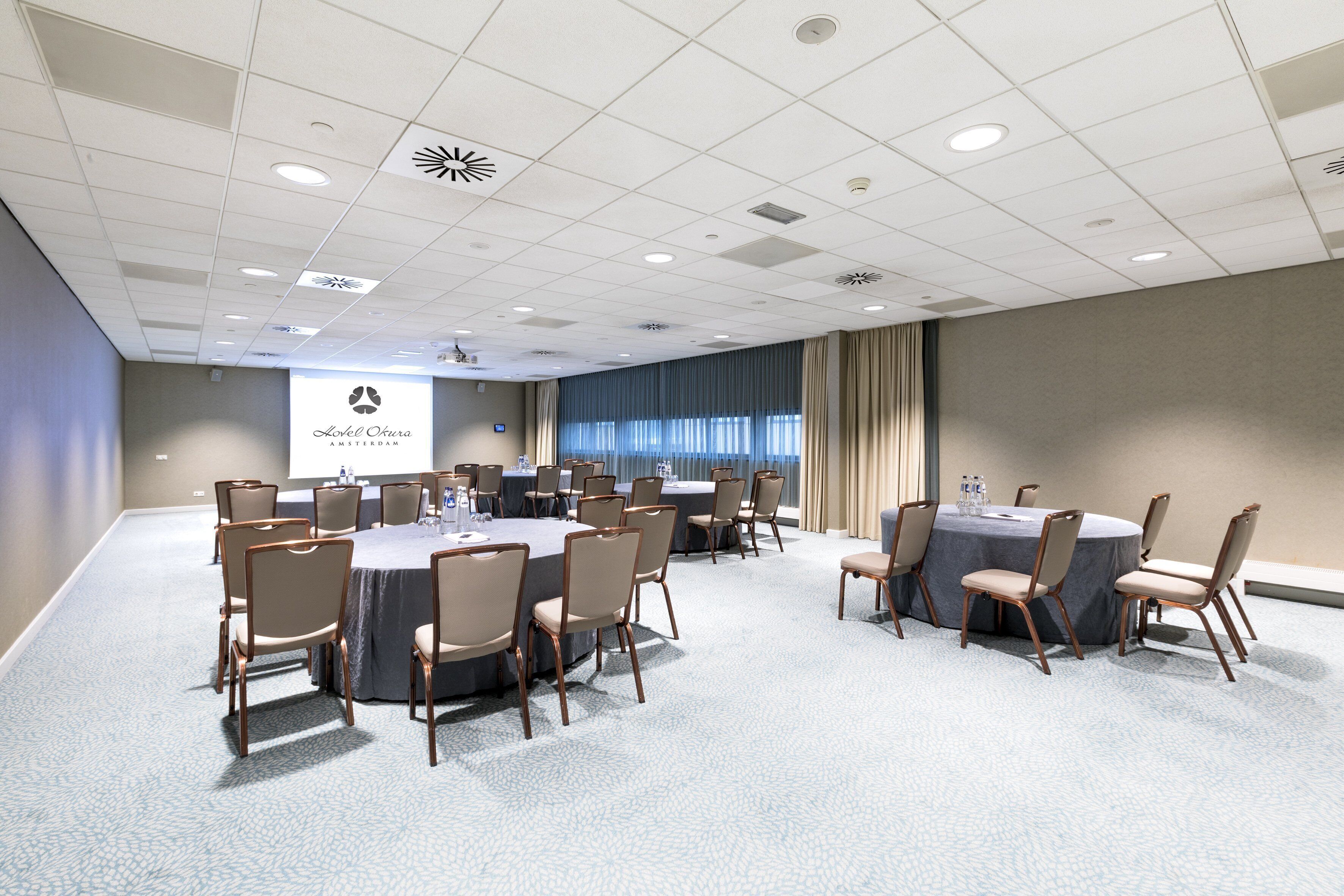 A conference room with round tables and chairs, a projector screen, and a view of the city through large windows.