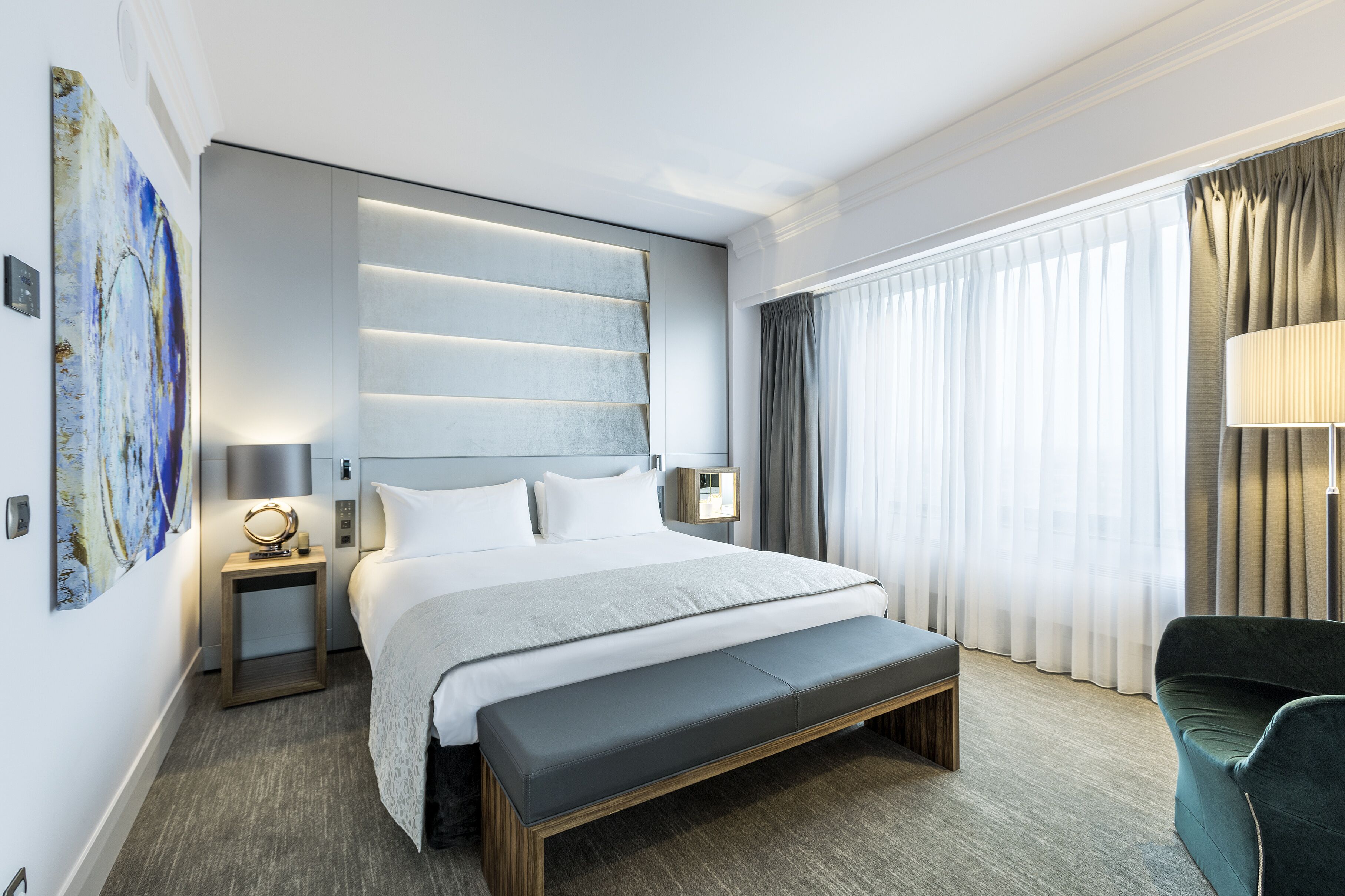 Una habitación de hotel moderna con una cama grande, un banco, una mesita de noche con lámpara, un panel de luz en la pared y un amplio ventanal con cortinas.