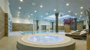 Un espace piscine intérieur moderne, doté d’un grand jacuzzi central, de coins salon confortables et d’une vue sur la piscine.