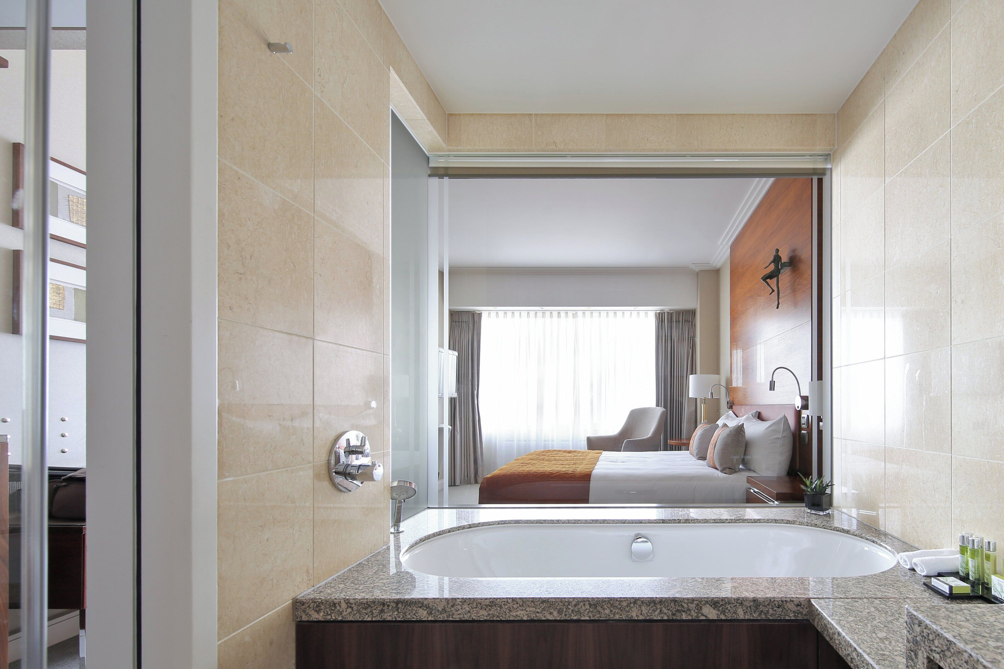 A modern hotel room with a large bathtub, a bed with a view, and a bathroom with a large mirror.