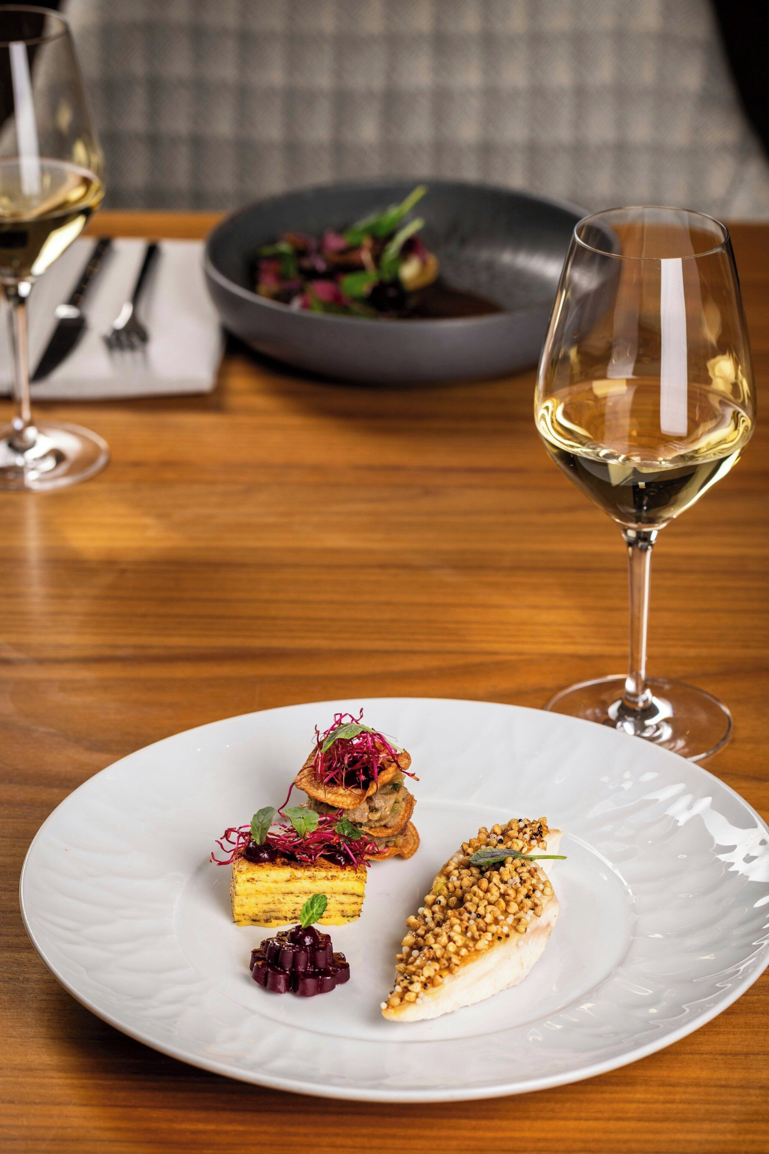 Una mesa de comedor con un plato blanco de comida, una copa de vino blanco y un bol de ensalada al fondo.