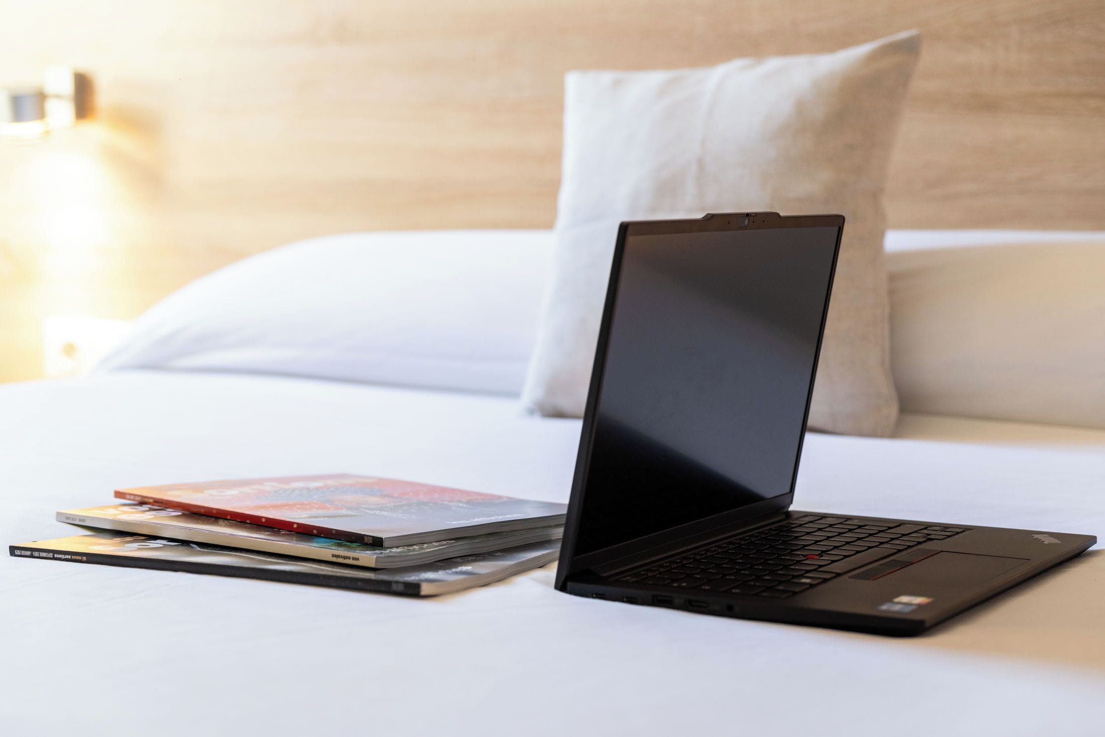 A laptop on a bed with a pillow and a lamp in the background.