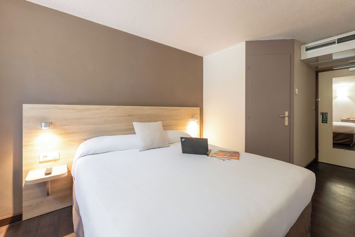 A hotel room with a large bed, bedside lamps, a laptop, and a door with a mirror.