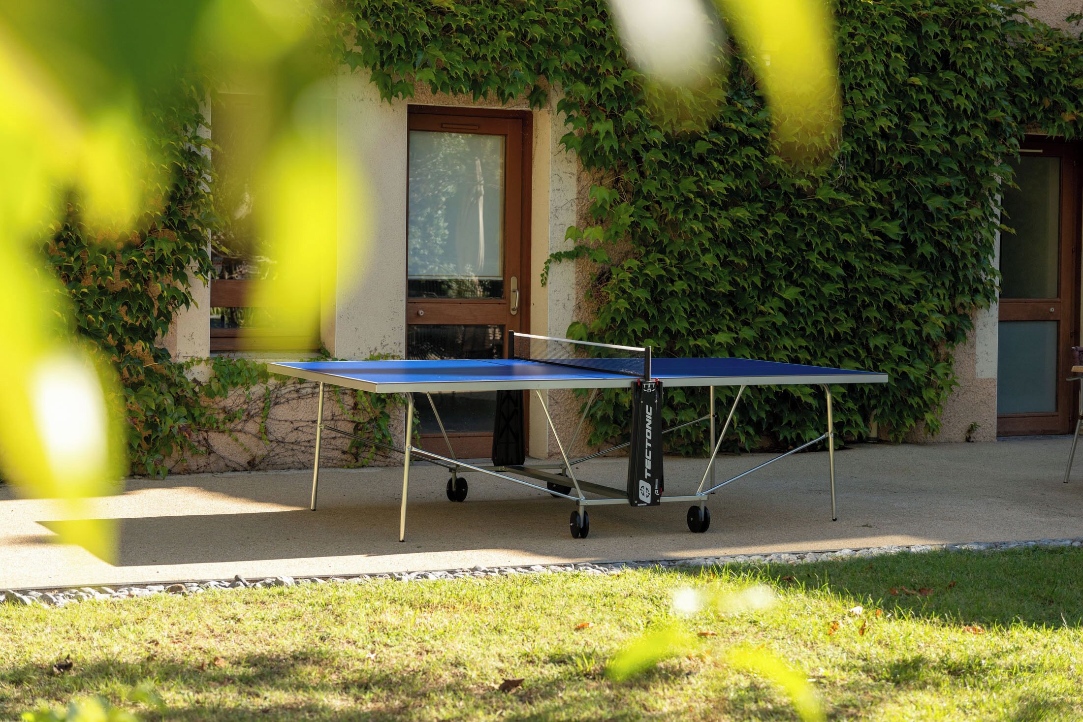 Une table de ping-pong installée dans un espace extérieur, avec un bâtiment recouvert de lierre en arrière-plan.