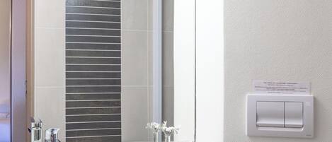 Un baño con lavamanos, espejo y retrete blancos. Hay una toalla colgada en la pared y un interruptor de luz.