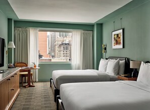 A hotel room with two beds, a desk, a television, and a view of the cityscape.