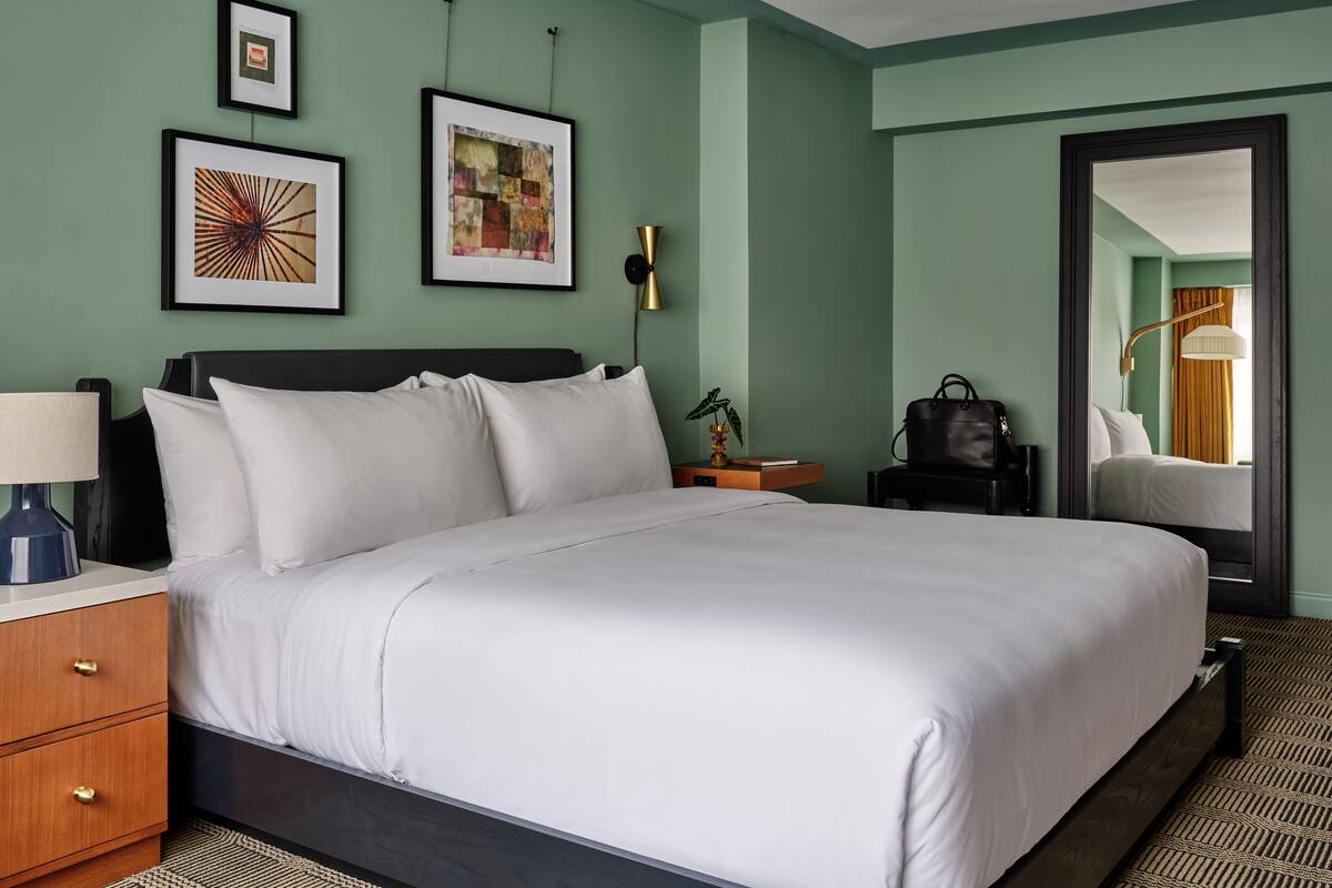 A hotel room with a large bed, a nightstand, a lamp, a mirror, and two framed artworks on the wall.