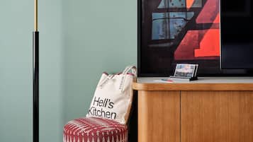 A modern hotel room with a floor lamp, a television, a cabinet, a patterned ottoman, and a tote bag.