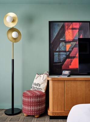 A modern hotel room with a floor lamp, a television, a cabinet, a patterned ottoman, and a tote bag.