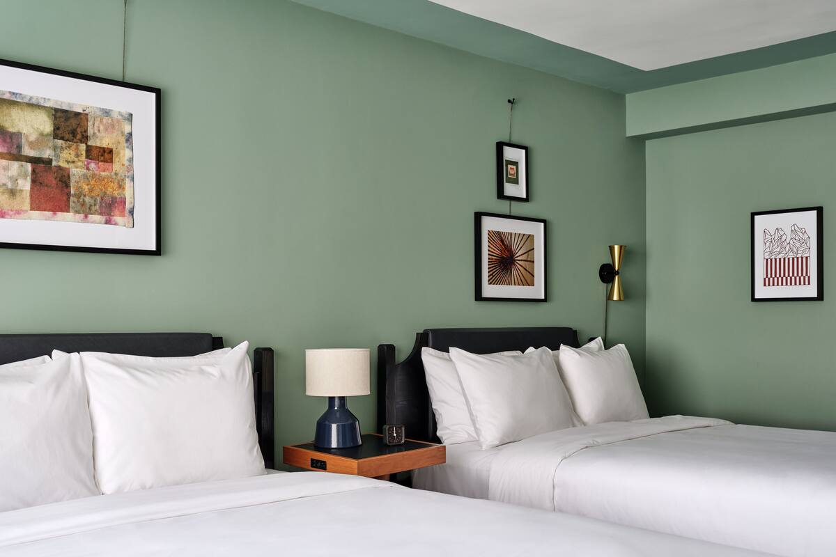 A hotel room with two beds, a nightstand with a lamp, and three framed artworks on the wall.