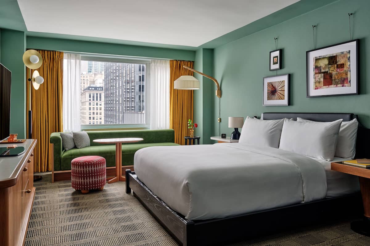 A hotel room with a large bed, a green sofa, a small table, and a view of the city through a window.