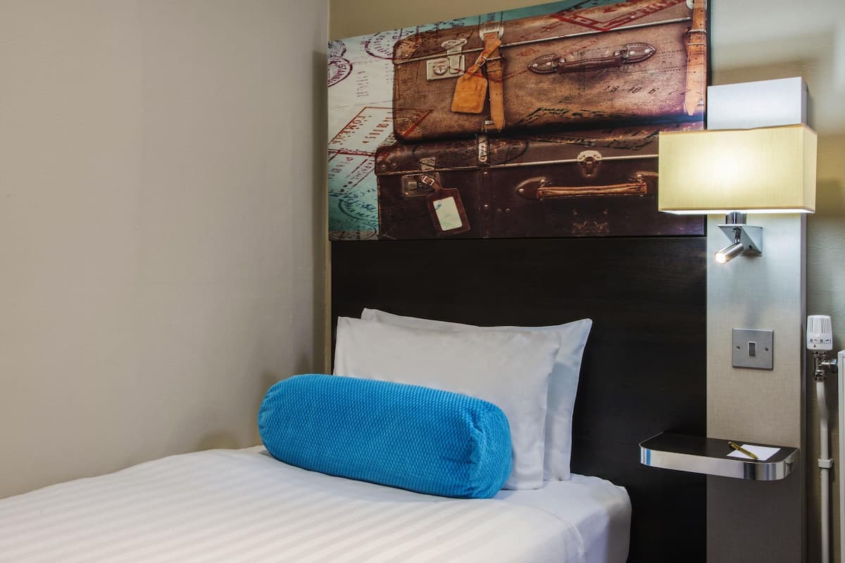 A hotel room with a bed, a headboard with a vintage suitcase design, a bedside lamp, and a nightstand.